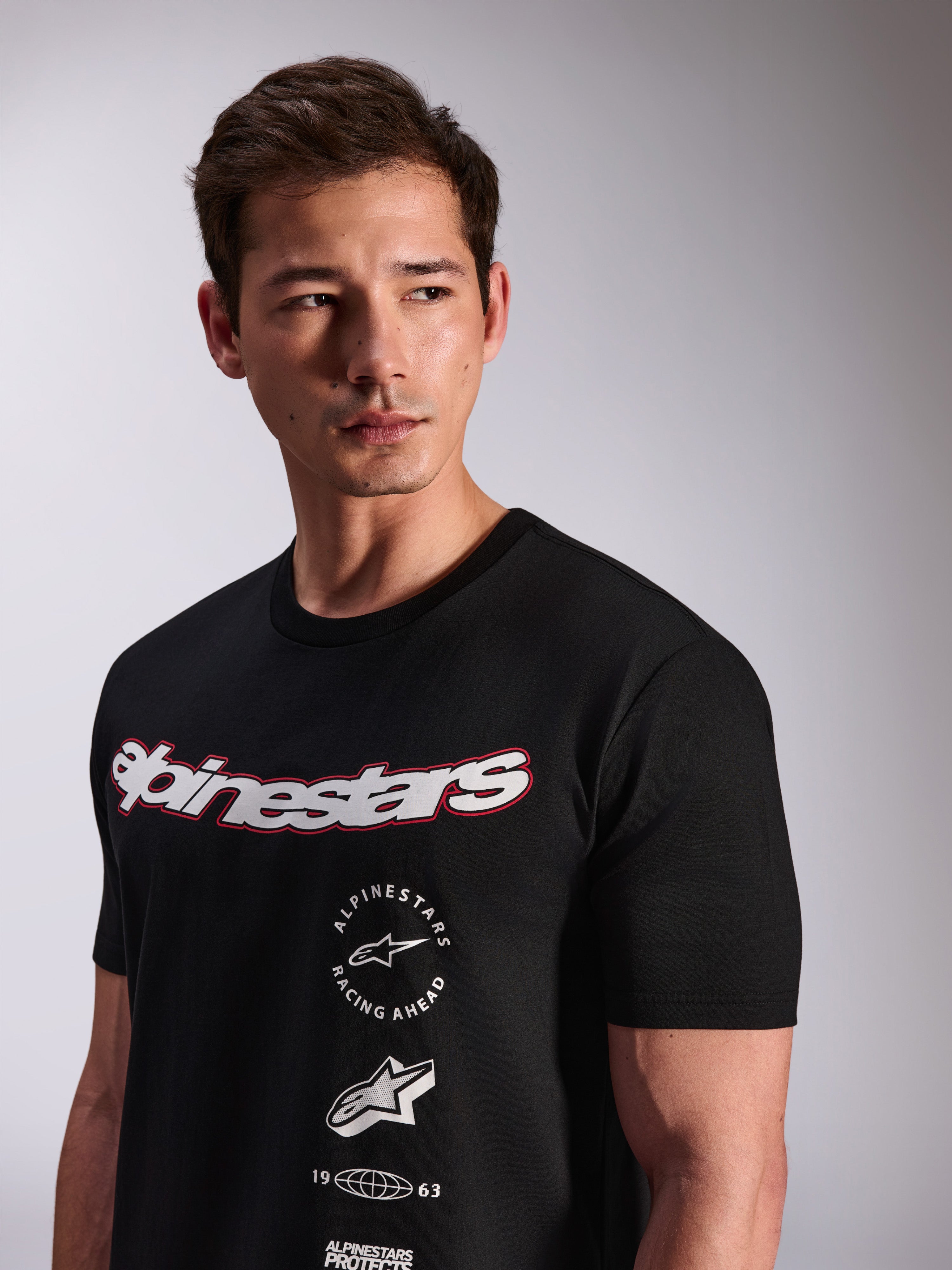 Alpinestars Abus CSF Tee, czarna koszulka z krótkim rękawem, noszona przez modela, białe logo Alpinestars z czerwoną obwódką i wielowarstwowe logotypy na klatce piersiowej, codzienna odzież streetwearowa w stylu motorsportowym