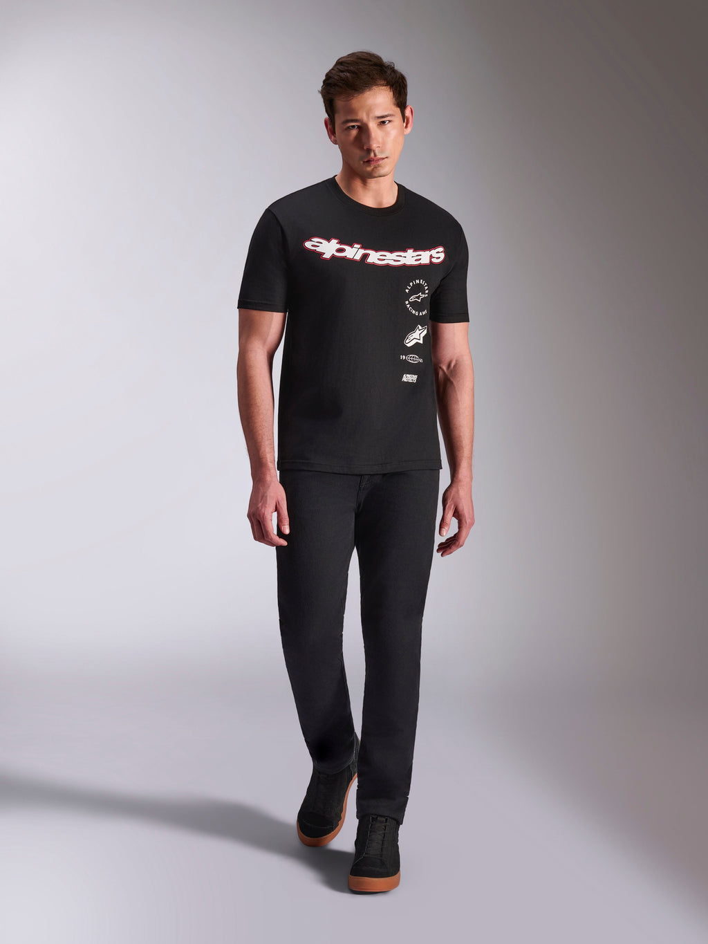 Alpinestars Abus CSF Tee, koszulka z krótkim rękawem, czarna, zakrzywiony czerwono-biały logotyp z wielowarstwowymi grafikami, noszona przez modela w czarnych spodniach i czarnych sneakersach, casualowy styl motorsportowy
