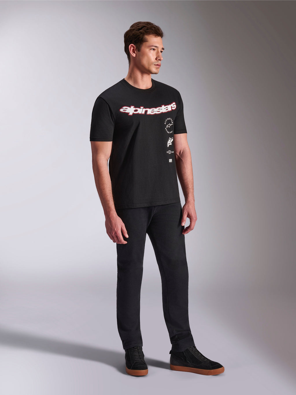 Alpinestars Abus CSF Tee, koszulka z krótkim rękawem, czarna, z zakrzywionym czerwono-białym logo Alpinestars i pionowo ułożonymi grafikami, noszona z czarnymi spodniami i sneakersami, casualowa odzież uliczna inspirowana motorsportem