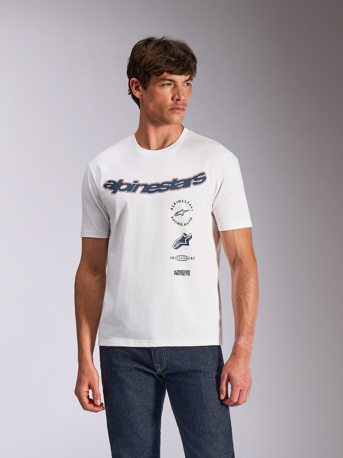 Alpinestars Abus CSF Tee, koszulka z krótkim rękawem, biała, zakrzywione logo Alpinestars z wielowarstwowymi grafikami na klatce piersiowej, model w ciemnych jeansach, casualowa odzież uliczna w stylu motorsport