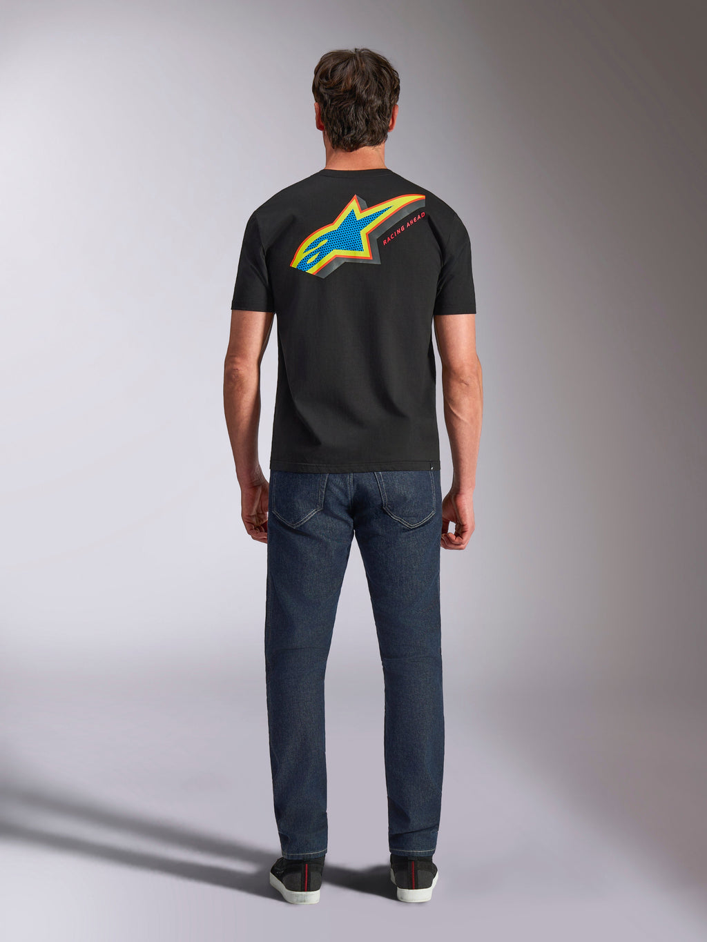 Koszulka Alpinestars Angler CSF Tee, t-shirt z krótkim rękawem, czarny, widok z tyłu na modela ubranego w koszulkę z dużym, wielokolorowym logo Astars i napisem „RACING AHEAD”, w połączeniu z ciemnoniebieskimi dżinsami, codzienna odzież uliczna w stylu motorsportowym