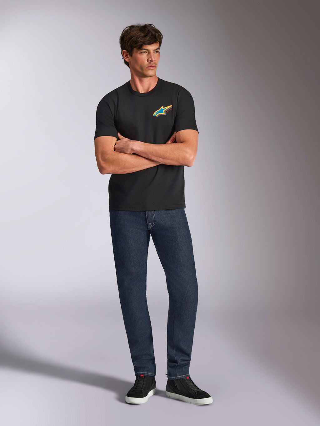 Alpinestars Angler CSF Tee, czarna koszulka z krótkim rękawem, żywe wielokolorowe logo Astars na piersi, model ze skrzyżowanymi ramionami, ciemne dżinsy i czarne sneakersy, casualowa odzież motorsportowa