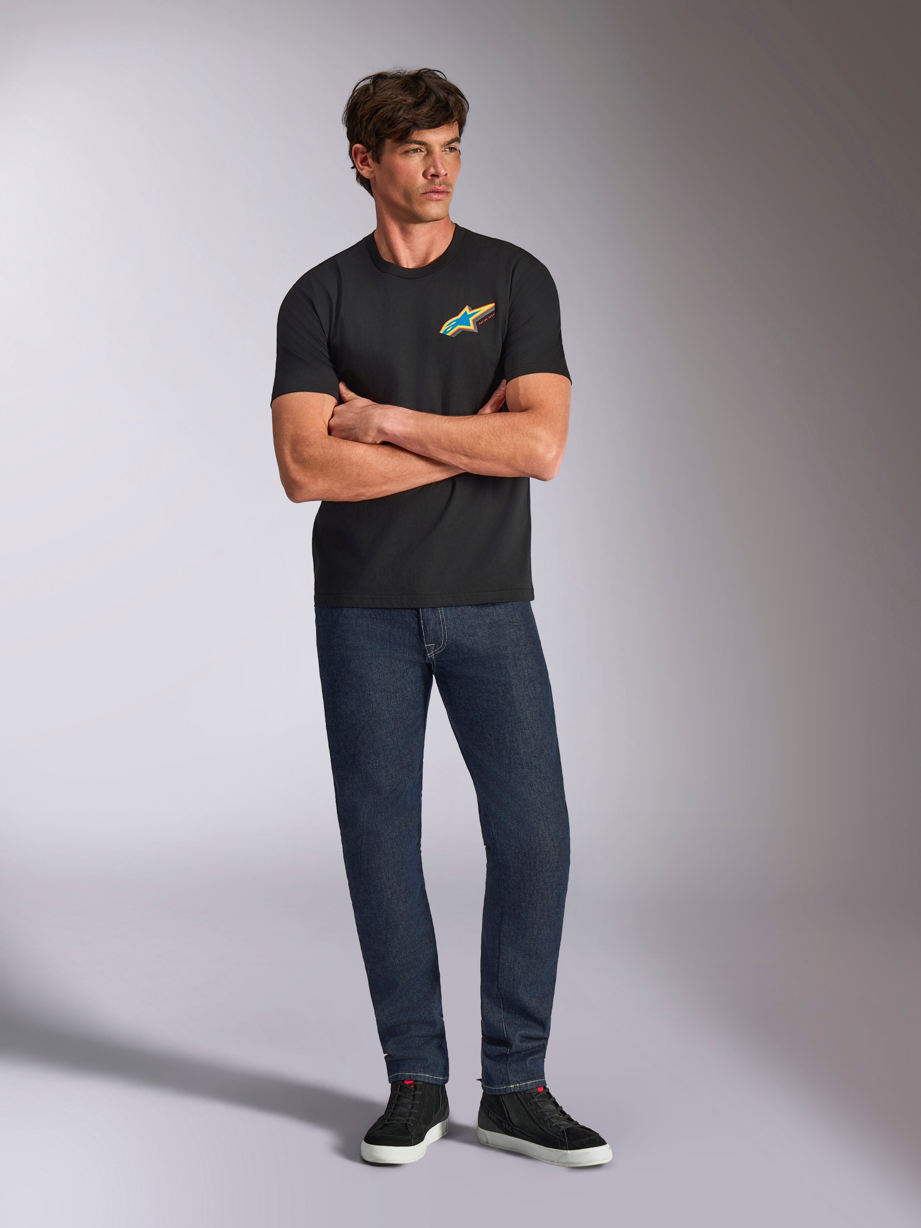 Alpinestars Angler CSF Tee, czarna koszulka z krótkim rękawem, żywe wielokolorowe logo Astars na piersi, model ze skrzyżowanymi ramionami, ciemne dżinsy i czarne sneakersy, casualowa odzież motorsportowa