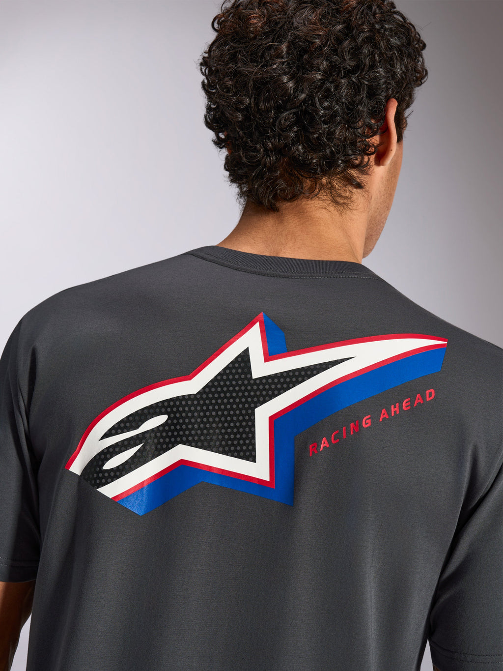 Alpinestars Angler CSF Tee, koszulka z krótkim rękawem, grafitowa, widok z tyłu na modelu z dużym wielokolorowym logo gwiazdy Alpinestars, perforowane wypełnienie i napis „RACING AHEAD”, codzienna odzież motorsportowa