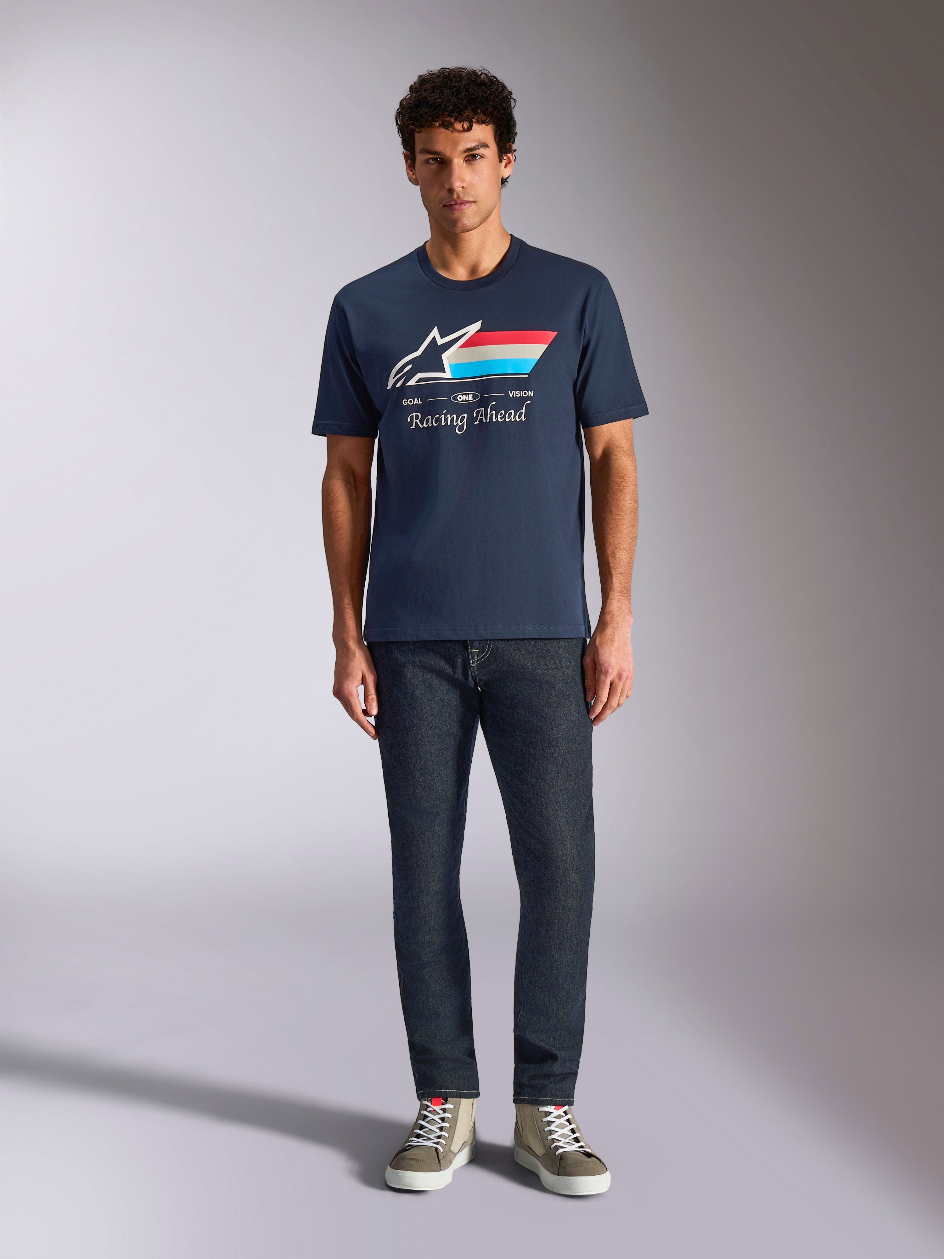 Alpinestars Trifecta CSF Tee, granatowa koszulka z krótkim rękawem, białe logo Astars z czerwonymi, białymi i jasnoniebieskimi pasami oraz napisem „Racing Ahead”, noszona z ciemnymi dżinsami i oliwkowymi sneakersami, codzienna odzież streetwearowa w stylu motorsportowym