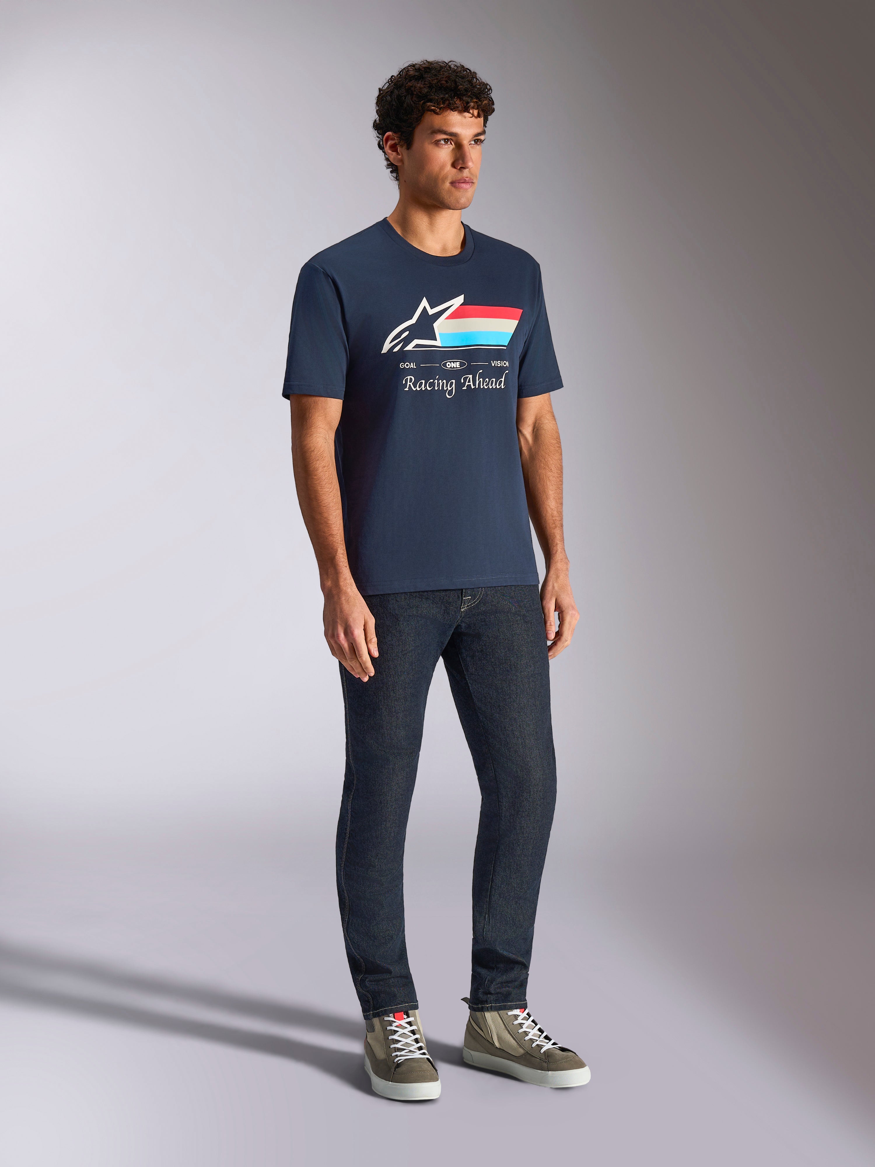 Koszulka Alpinestars Trifecta CSF, t-shirt z krótkim rękawem, granatowa, z logo Astars, trójkolorowymi paskami i napisem „Racing Ahead”, noszona przez modela z ciemnymi dżinsami i sneakersami, codzienna odzież uliczna inspirowana sportami motorowymi