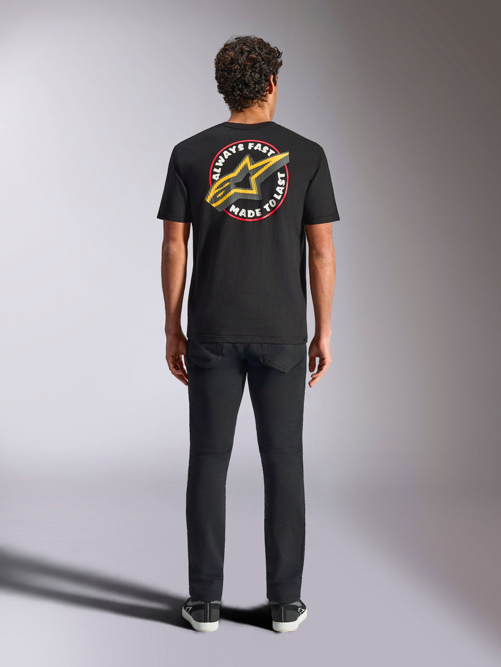 Alpinestars Aorbit CSF Tee, koszulka z krótkim rękawem, czarna, widok z tyłu na modelu w czarnych spodniach, z dużą grafiką „Always Fast Made to Last” i żółtym logo Astars, casualowa odzież uliczna w stylu motorsport