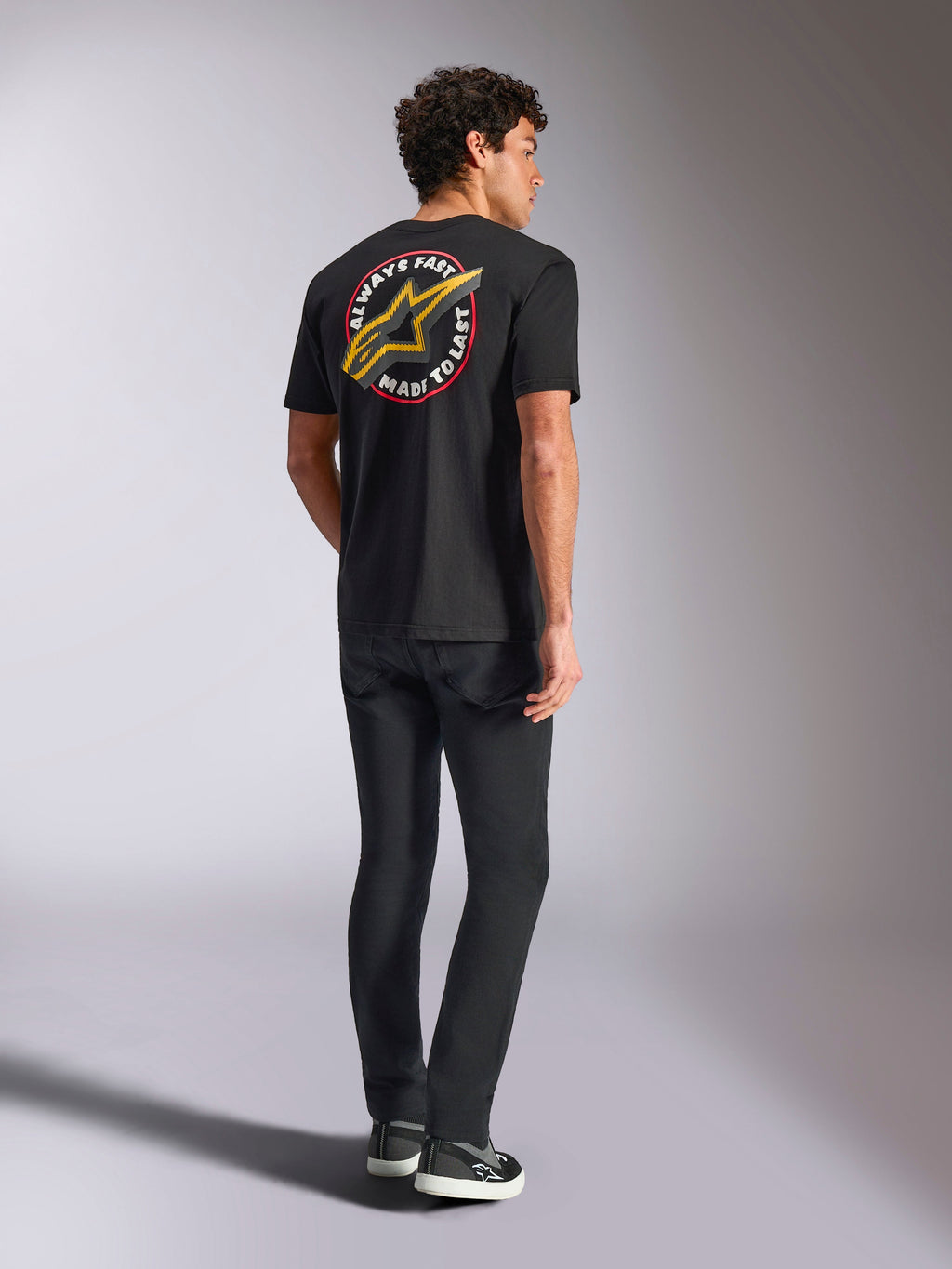 Alpinestars Aorbit CSF Tee, koszulka z krótkim rękawem, czarna, widok z tyłu z dużym żółtym logo Astars i okrągłą grafiką „Always Fast Made to Last”, zestawiona z czarnymi dżinsami i sneakersami, styl streetwear inspirowany motorsportem