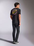 Alpinestars Aorbit CSF Tee, koszulka z krótkim rękawem, czarna, widok z tyłu z dużym żółtym logo Astars i okrągłą grafiką „Always Fast Made to Last”, zestawiona z czarnymi dżinsami i sneakersami, styl streetwear inspirowany motorsportem