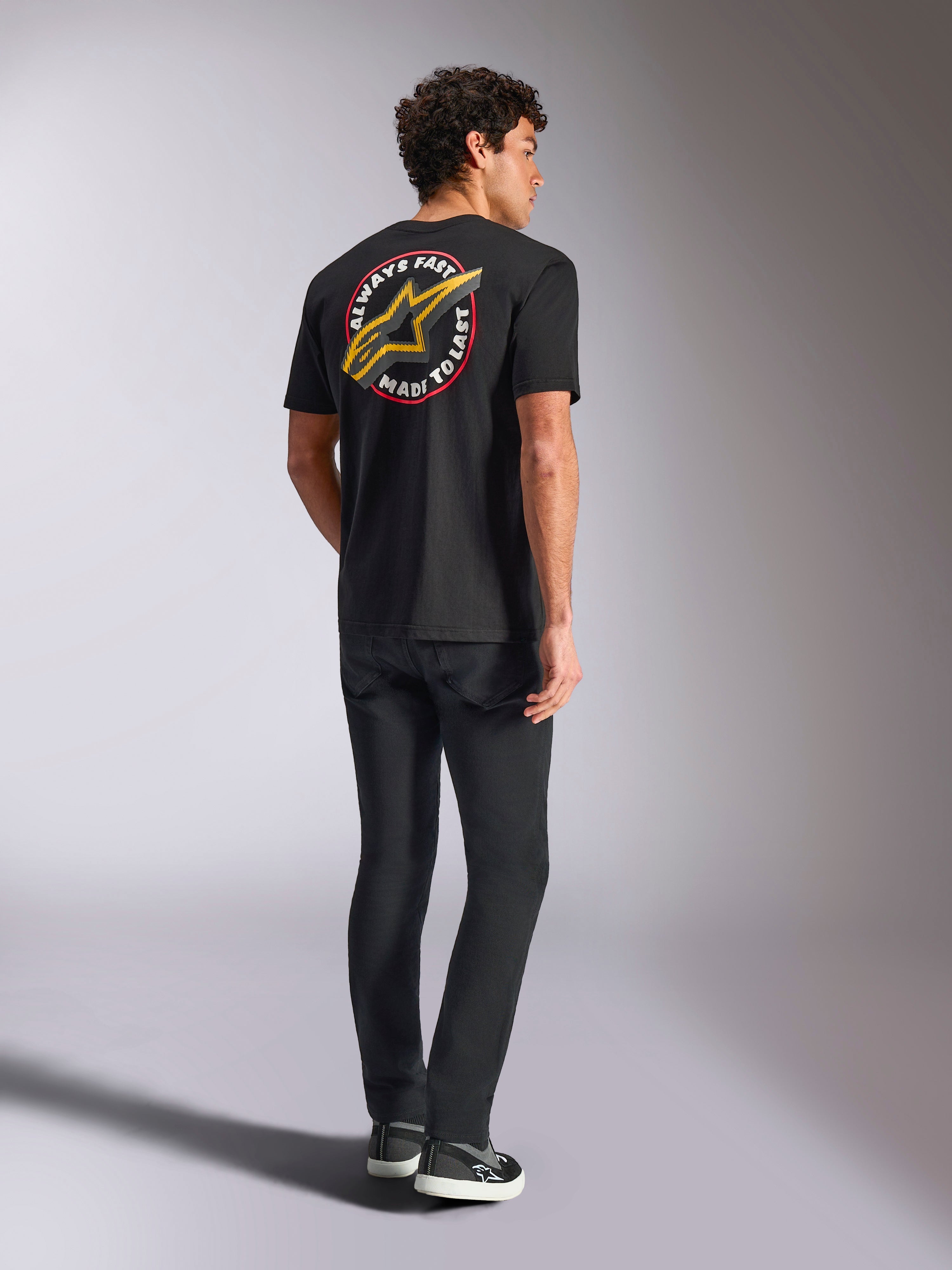 Alpinestars Aorbit CSF Tee, koszulka z krótkim rękawem, czarna, widok z tyłu z dużym żółtym logo Astars i okrągłą grafiką „Always Fast Made to Last”, zestawiona z czarnymi dżinsami i sneakersami, styl streetwear inspirowany motorsportem