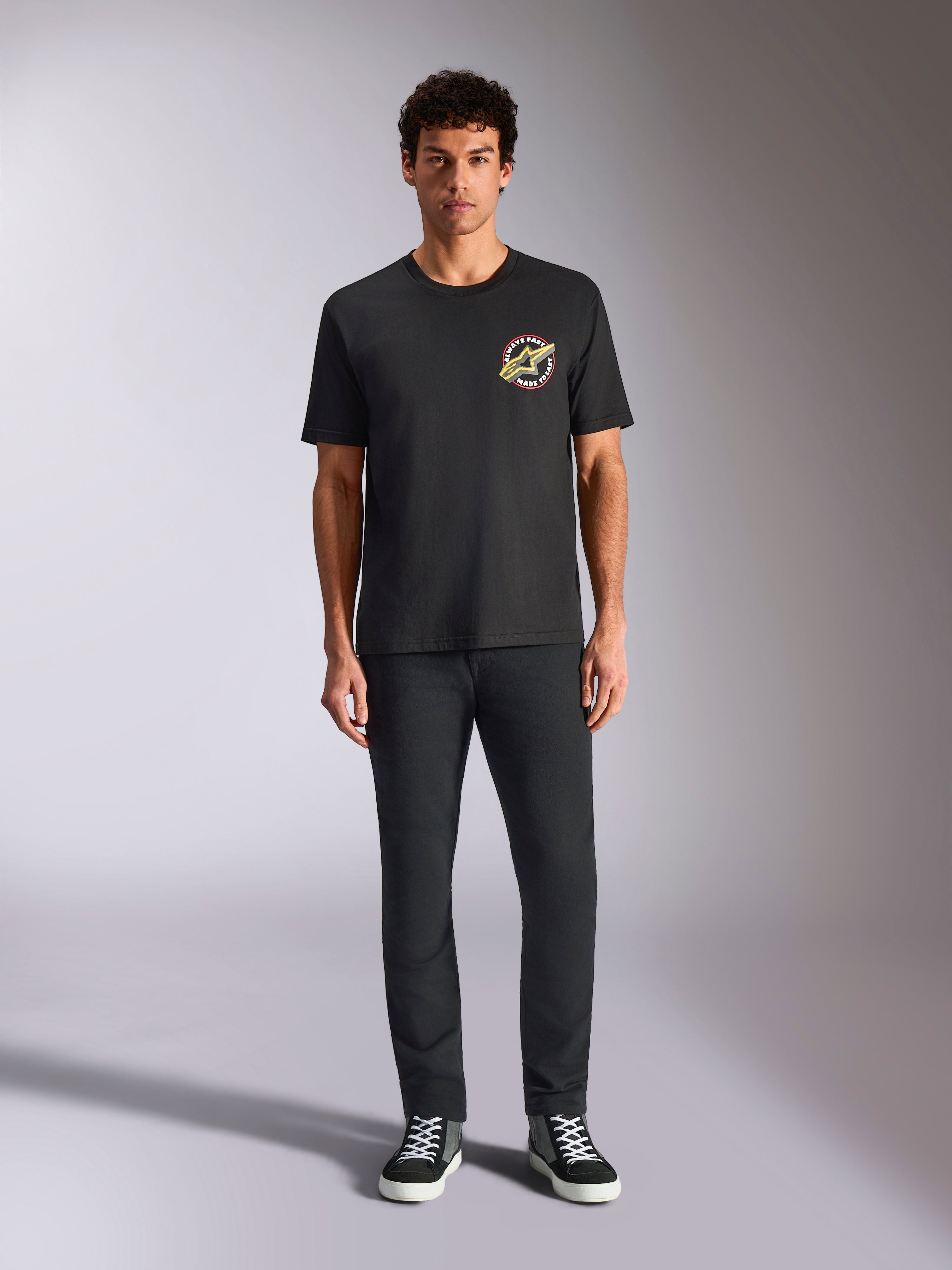 Alpinestars Aorbit CSF Tee, koszulka z krótkim rękawem, czarna, okrągłe logo z napisem „Always Fast Made to Last” na piersi, model w czarnych spodniach i sneakersach, codzienna odzież uliczna inspirowana sportami motorowymi