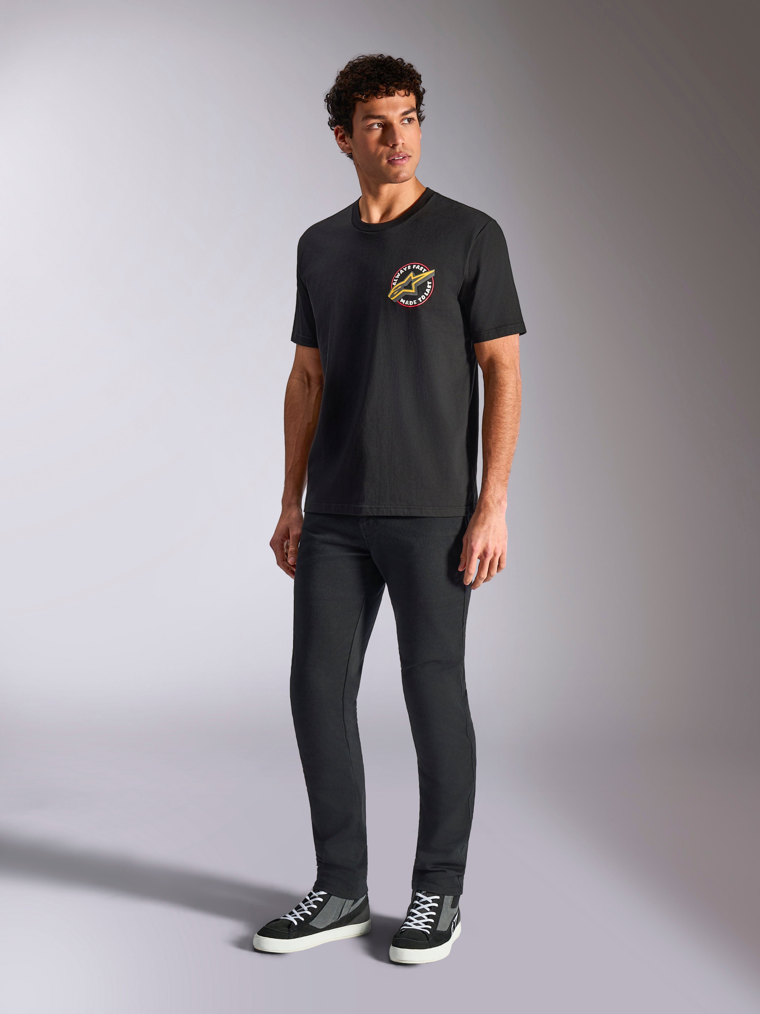 Alpinestars Aorbit CSF Tee, czarna koszulka z krótkim rękawem, okrągła grafika „Always Fast Made to Last” na piersi, model w ciemnych spodniach typu slim i sneakersach, casualowy styl motorsport