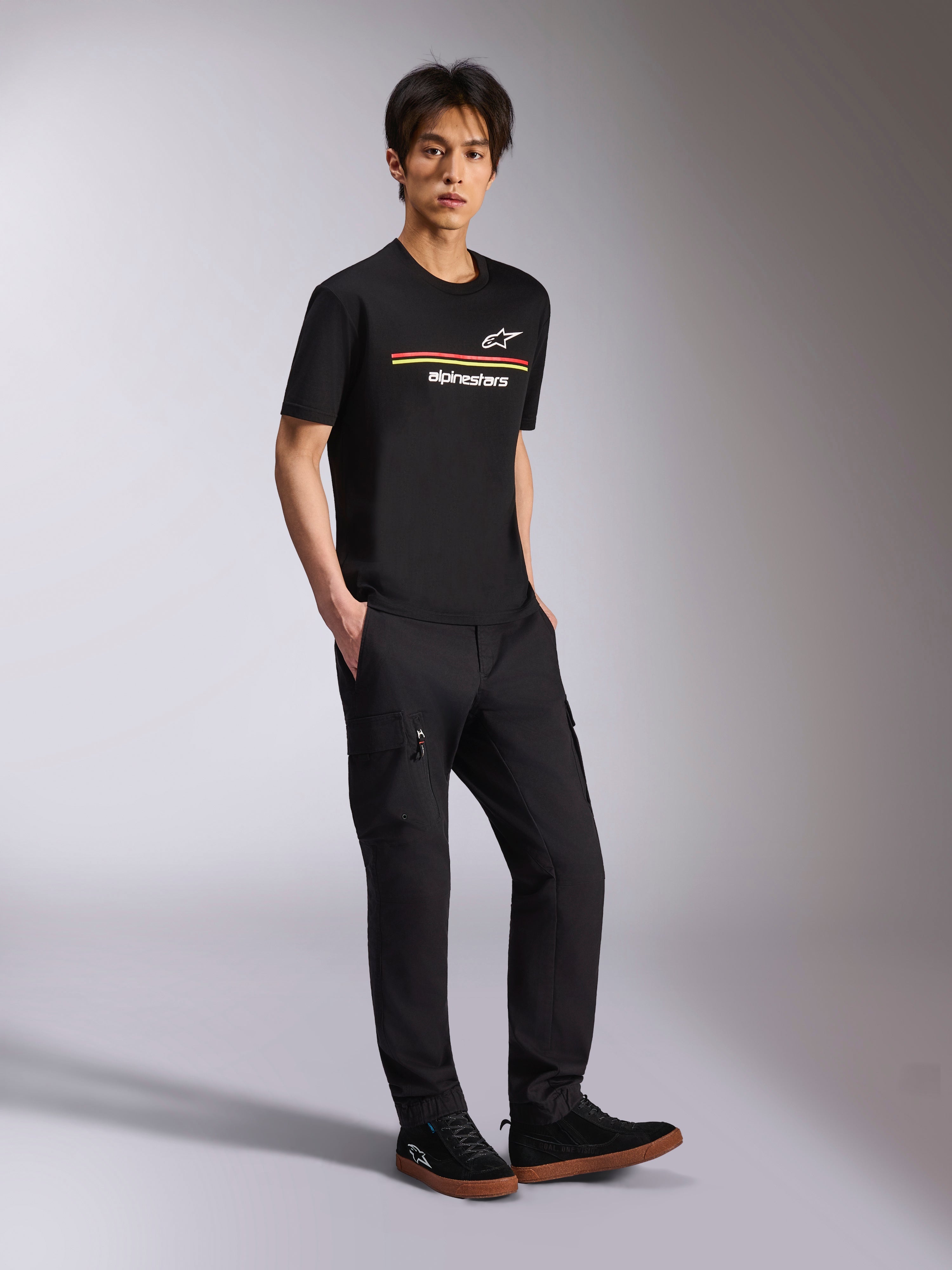 Alpinestars Lanes CSF Tee, czarna koszulka z krótkim rękawem, kolorowe logo Astars i paski Alpinestars na klatce piersiowej, model ubrany w czarne spodnie cargo, casualowy styl motorsportowy