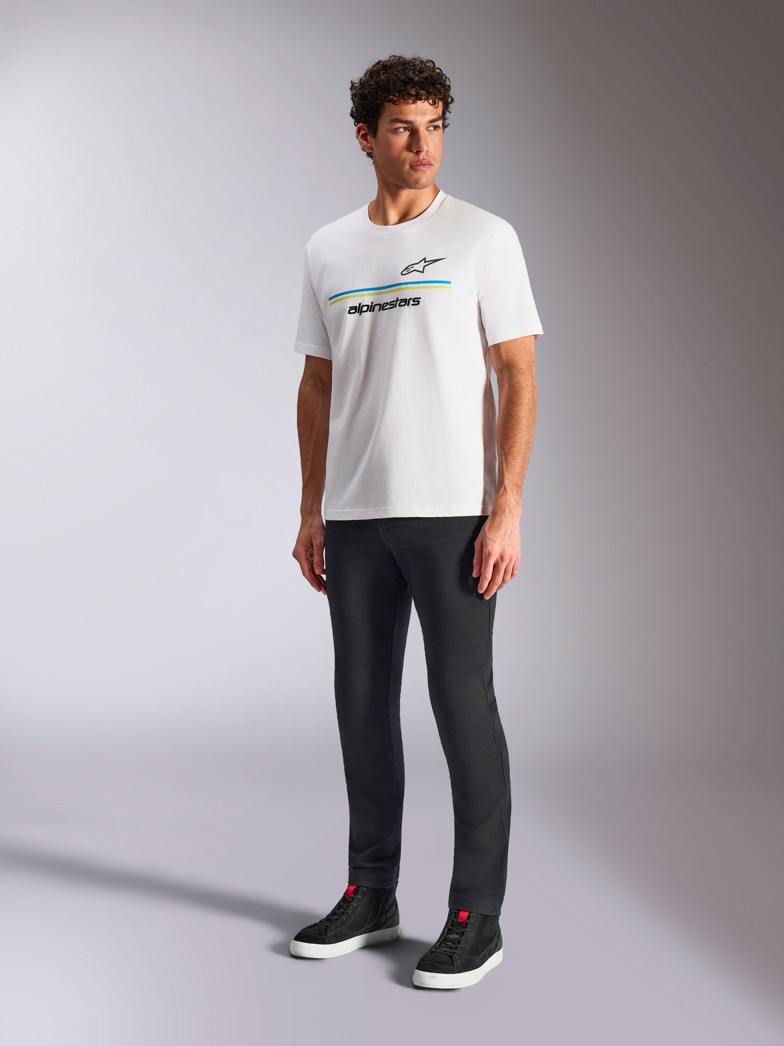Alpinestars Lanes CSF Tee, biała koszulka z krótkim rękawem, model w czarnych spodniach slim-fit i czarnych wysokich sneakersach, z niebieskimi i żółtymi poziomymi paskami oraz logo Alpinestars na piersi, codzienna odzież streetwearowa inspirowana sportami motorowymi