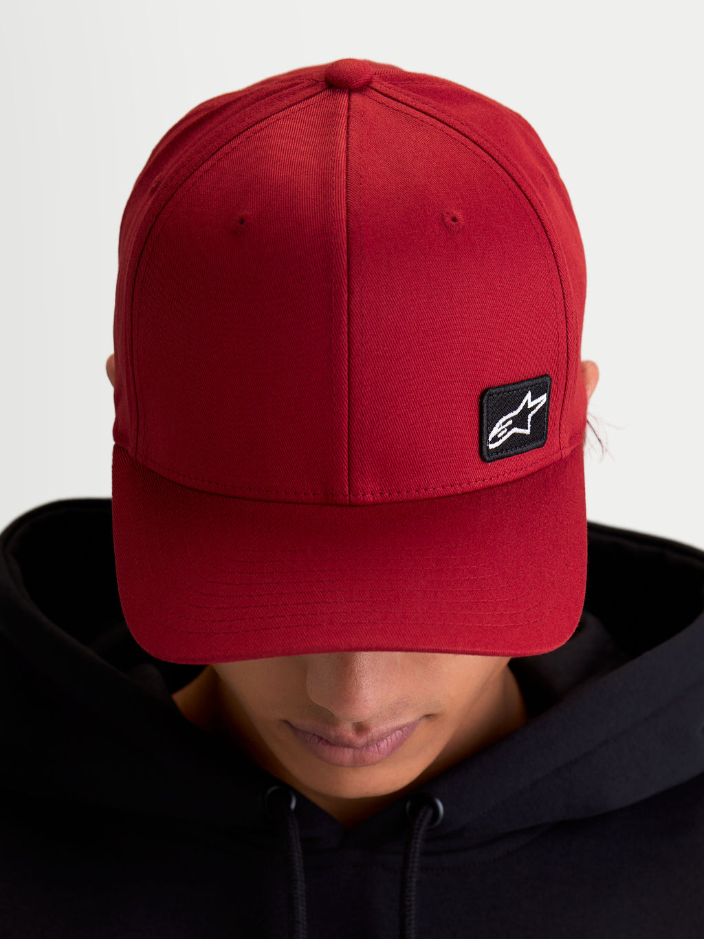 Alpinestars czapka Pivot, sportowa czapka z daszkiem typu baseball cap, ciemnoczerwona, unisex, zakrzywiony daszek i usztywniona korona, mała czarna haftowana naszywka z logo Astars na trwałej tkaninie keprowej, widok na modelu.