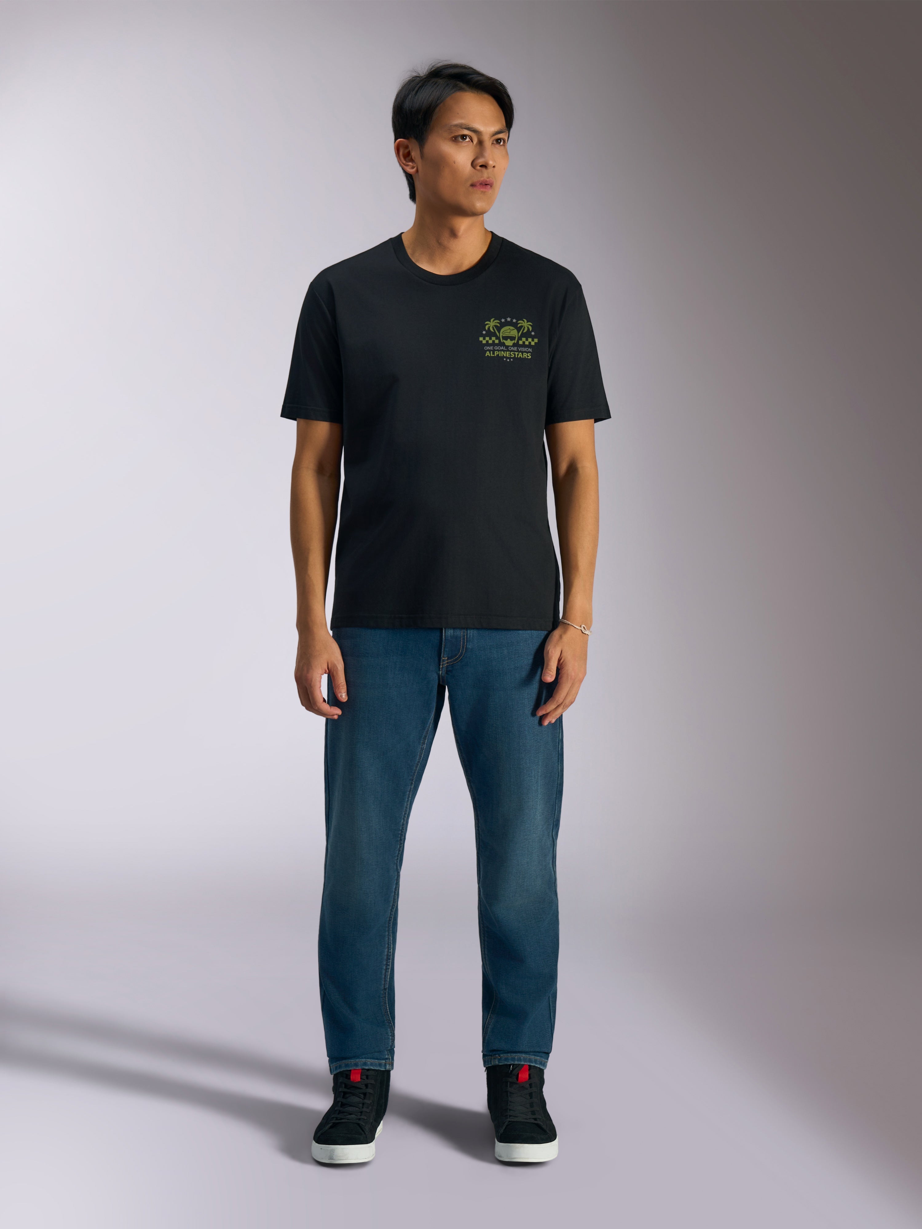 Alpinestars Order CSF Tee, czarna koszulka z krótkim rękawem, limonkowa grafika z kaskiem i palmami na piersi, zestawiona z niebieskimi jeansami i czarnymi sneakersami, codzienna odzież motorsportowa
