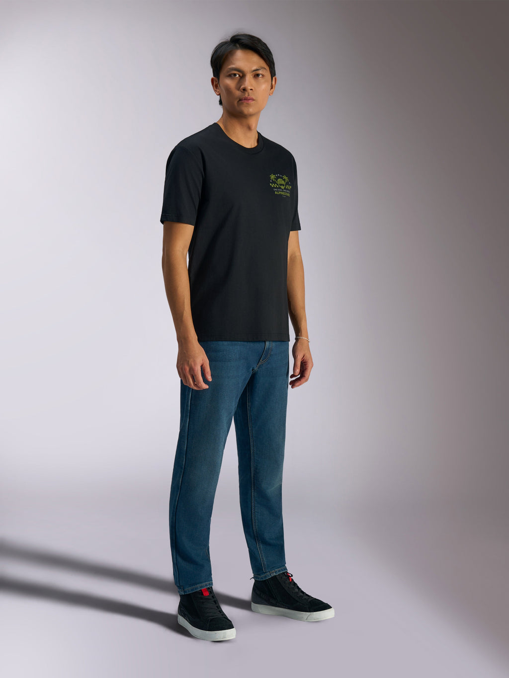 Alpinestars Order CSF Tee, czarna koszulka z krótkim rękawem, zielone logo z hełmem i palmami na piersi, codzienna odzież motorsportowa, noszona z niebieskimi dżinsami i czarnymi sneakersami