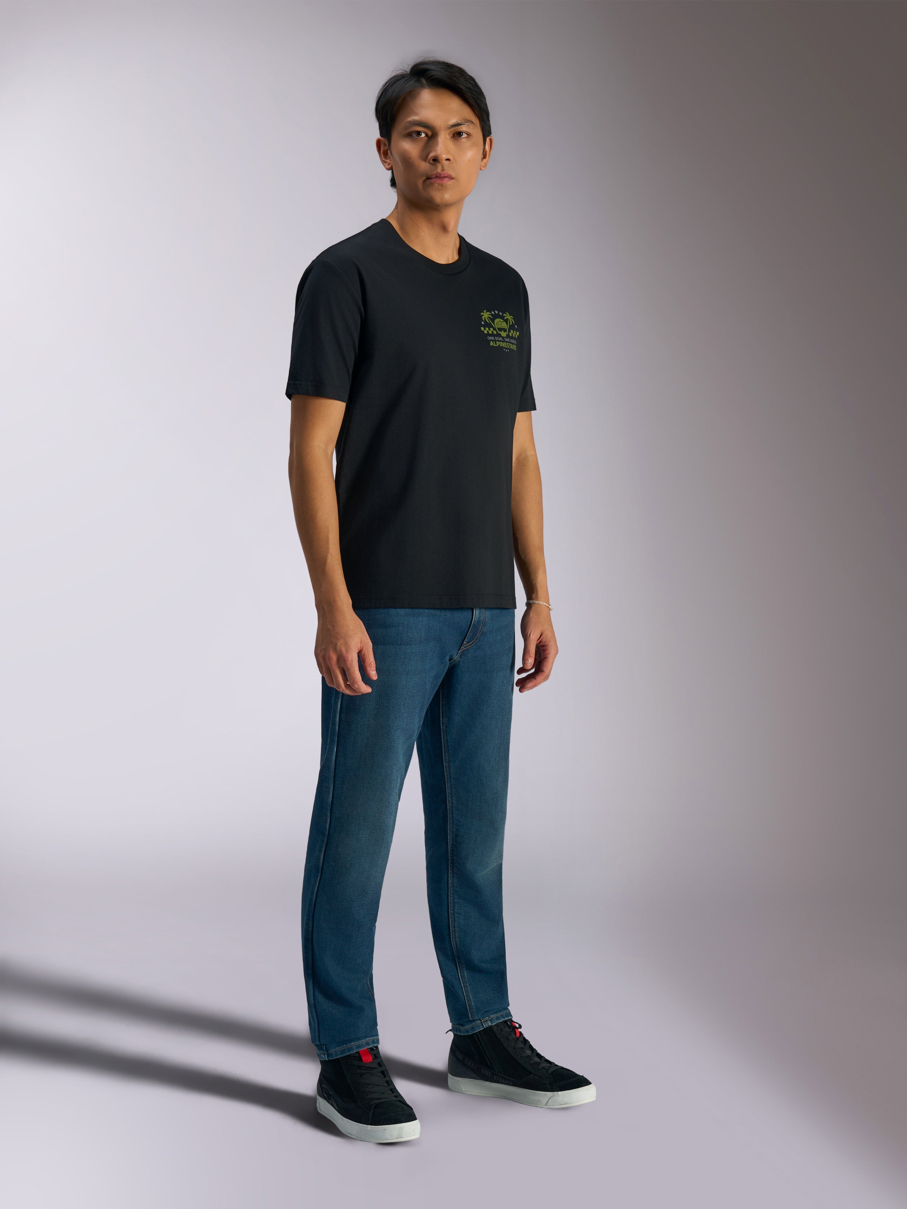 Alpinestars Order CSF Tee, czarna koszulka z krótkim rękawem, zielone logo z hełmem i palmami na piersi, codzienna odzież motorsportowa, noszona z niebieskimi dżinsami i czarnymi sneakersami