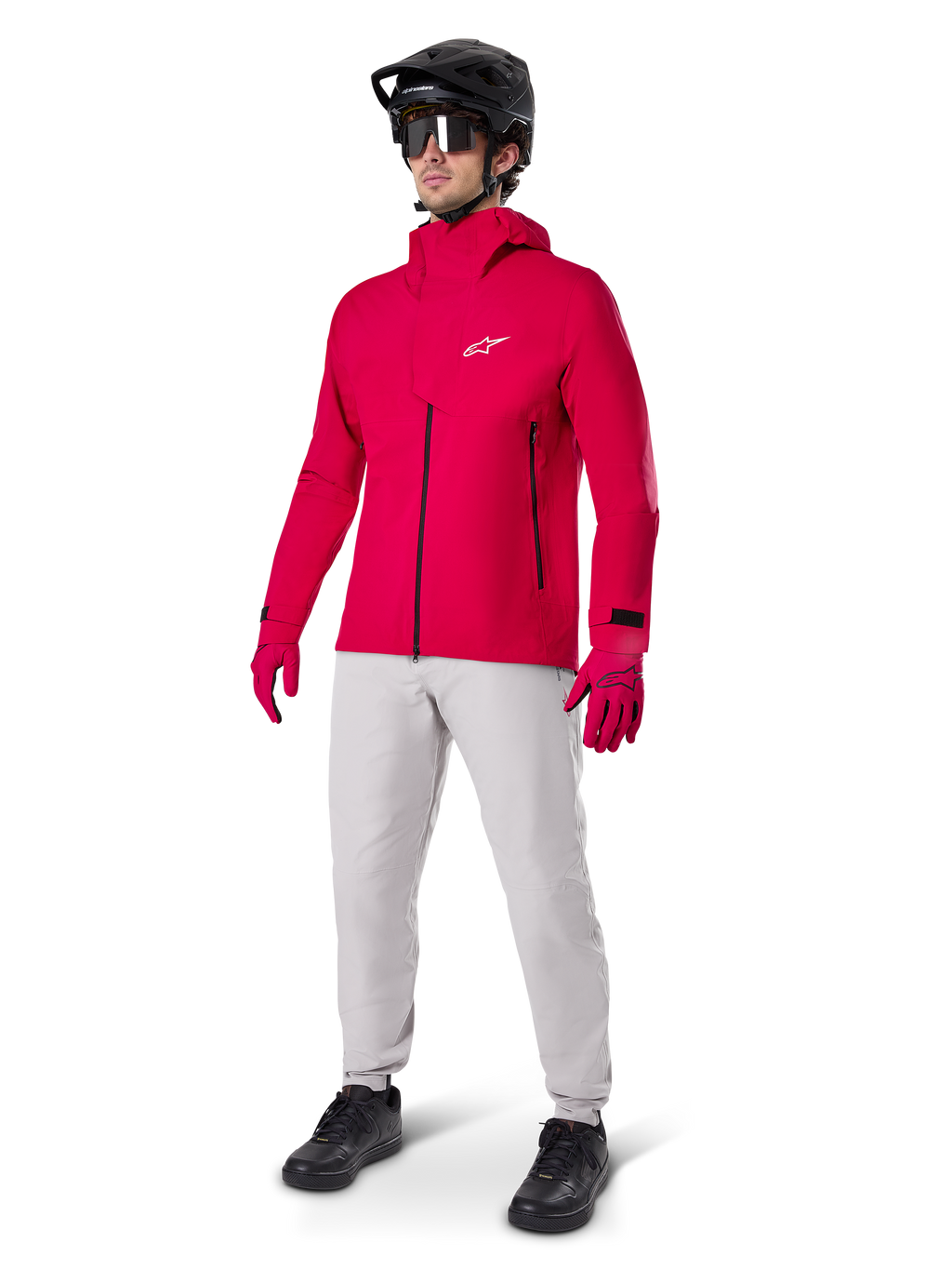 Rowerzysta ubrany w wodoodporną kurtkę Alpinestars A-Dura Elite w kolorze Red Fluo, jasnoszare spodnie techniczne, czerwone rękawiczki, czarny kask MTB z goglami i czarne buty; kompletny, wysokowydajny strój do kolarstwa górskiego.