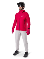 Rowerzysta ubrany w wodoodporną kurtkę Alpinestars A-Dura Elite w kolorze Red Fluo, jasnoszare spodnie techniczne, czerwone rękawiczki, czarny kask MTB z goglami i czarne buty; kompletny, wysokowydajny strój do kolarstwa górskiego.