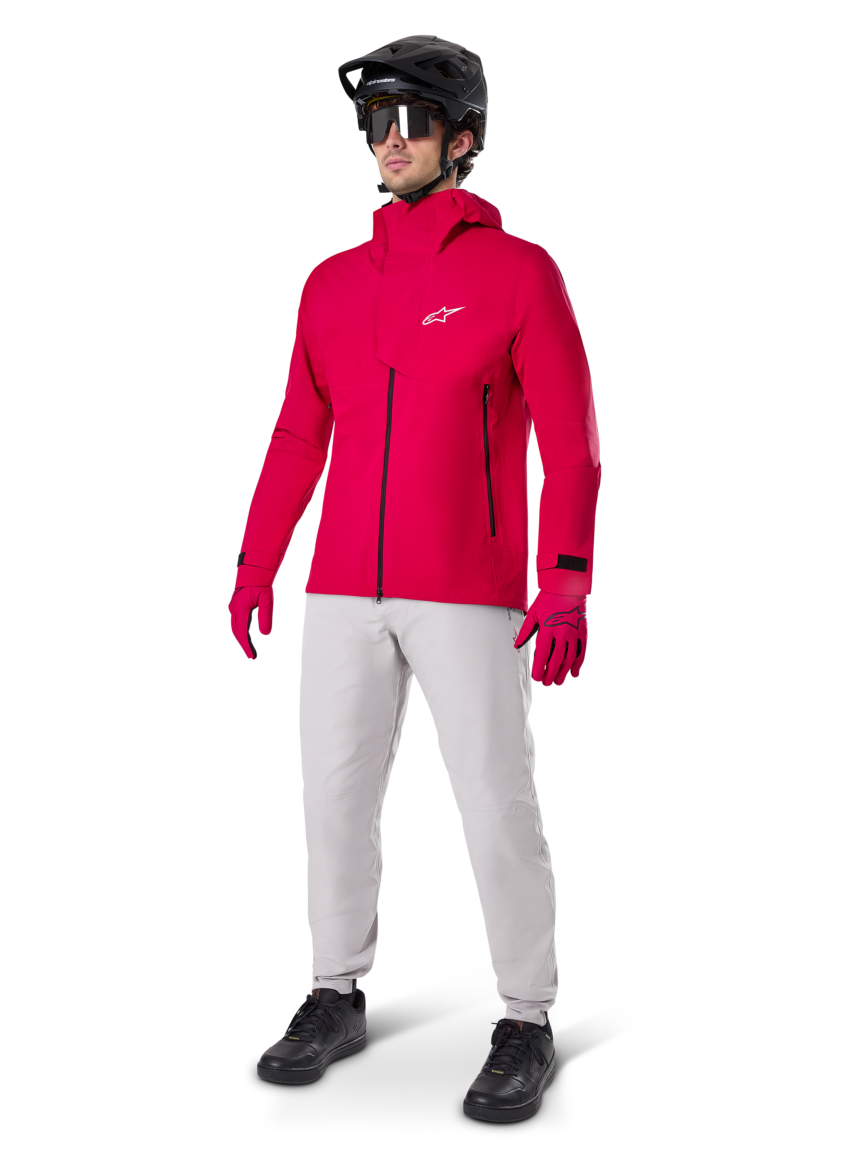 Rowerzysta ubrany w wodoodporną kurtkę Alpinestars A-Dura Elite w kolorze Red Fluo, jasnoszare spodnie techniczne, czerwone rękawiczki, czarny kask MTB z goglami i czarne buty; kompletny, wysokowydajny strój do kolarstwa górskiego.