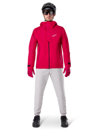 Wodoodporna kurtka Alpinestars A-Dura Elite, Red Fluo, techniczna kurtka rowerowa MTB noszona z czarnym kaskiem MTB, okularami przeciwsłonecznymi, pasującymi czerwonymi rękawiczkami i jasnoszarymi spodniami, widok całej sylwetki w odzieży rowerowej na każdą pogodę