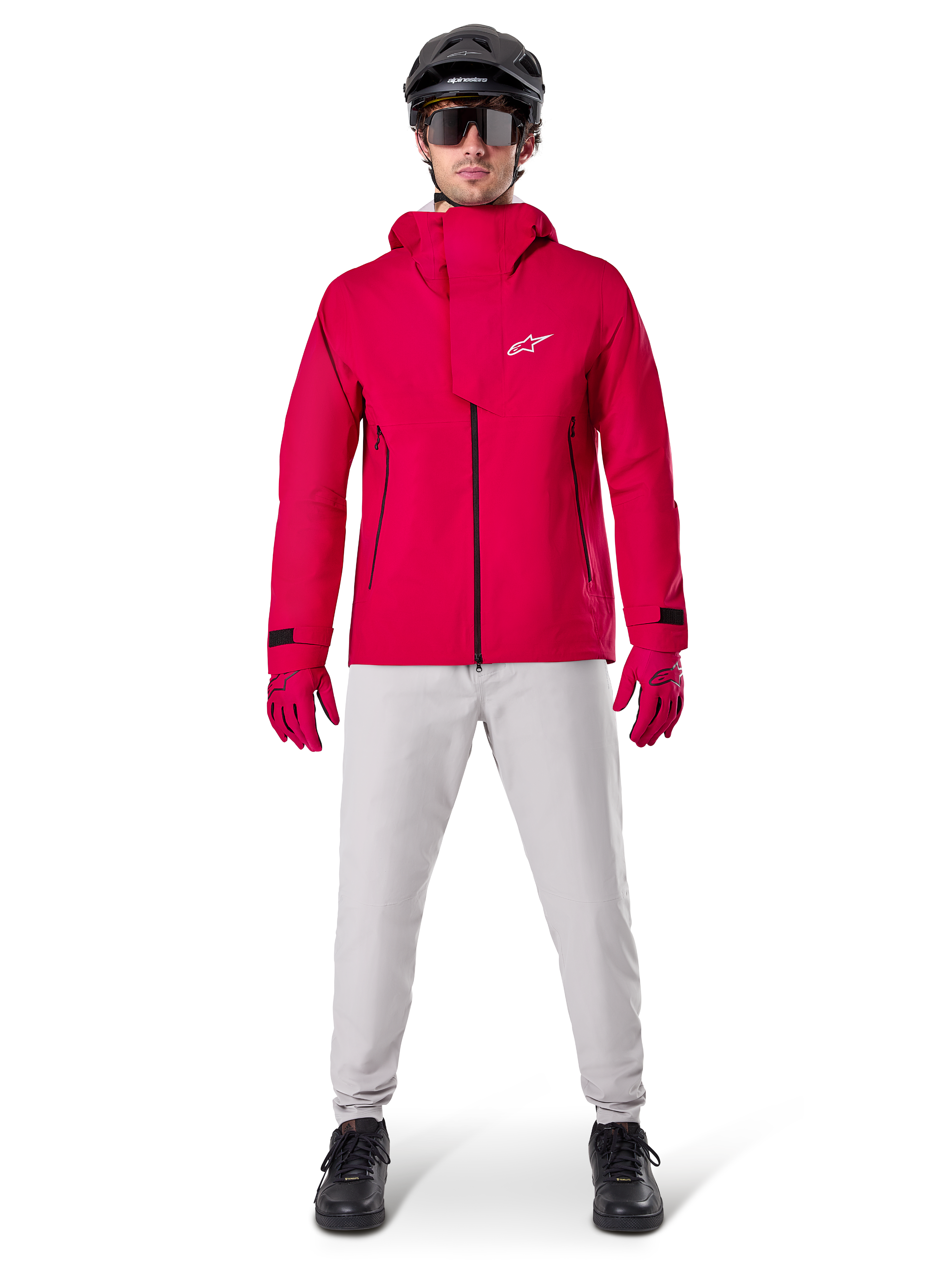Wodoodporna kurtka Alpinestars A-Dura Elite, Red Fluo, techniczna kurtka rowerowa MTB noszona z czarnym kaskiem MTB, okularami przeciwsłonecznymi, pasującymi czerwonymi rękawiczkami i jasnoszarymi spodniami, widok całej sylwetki w odzieży rowerowej na każdą pogodę