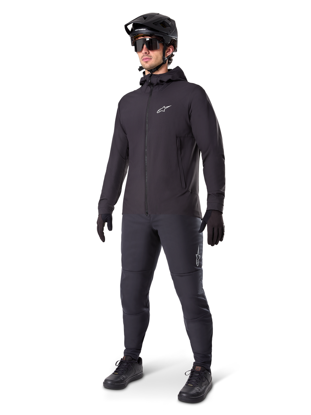 Alpinestars A-Dura Thermal Jacket w kolorze czarnym, noszona przez rowerzystę w pełnym stroju MTB, w tym czarnym kasku, okularach przeciwsłonecznych i spodniach rowerowych, lekka ocieplana kurtka z kapturem do jazdy w niskich temperaturach.