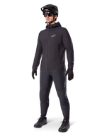 Alpinestars A-Dura Thermal Jacket w kolorze czarnym, noszona przez rowerzystę w pełnym stroju MTB, w tym czarnym kasku, okularach przeciwsłonecznych i spodniach rowerowych, lekka ocieplana kurtka z kapturem do jazdy w niskich temperaturach.