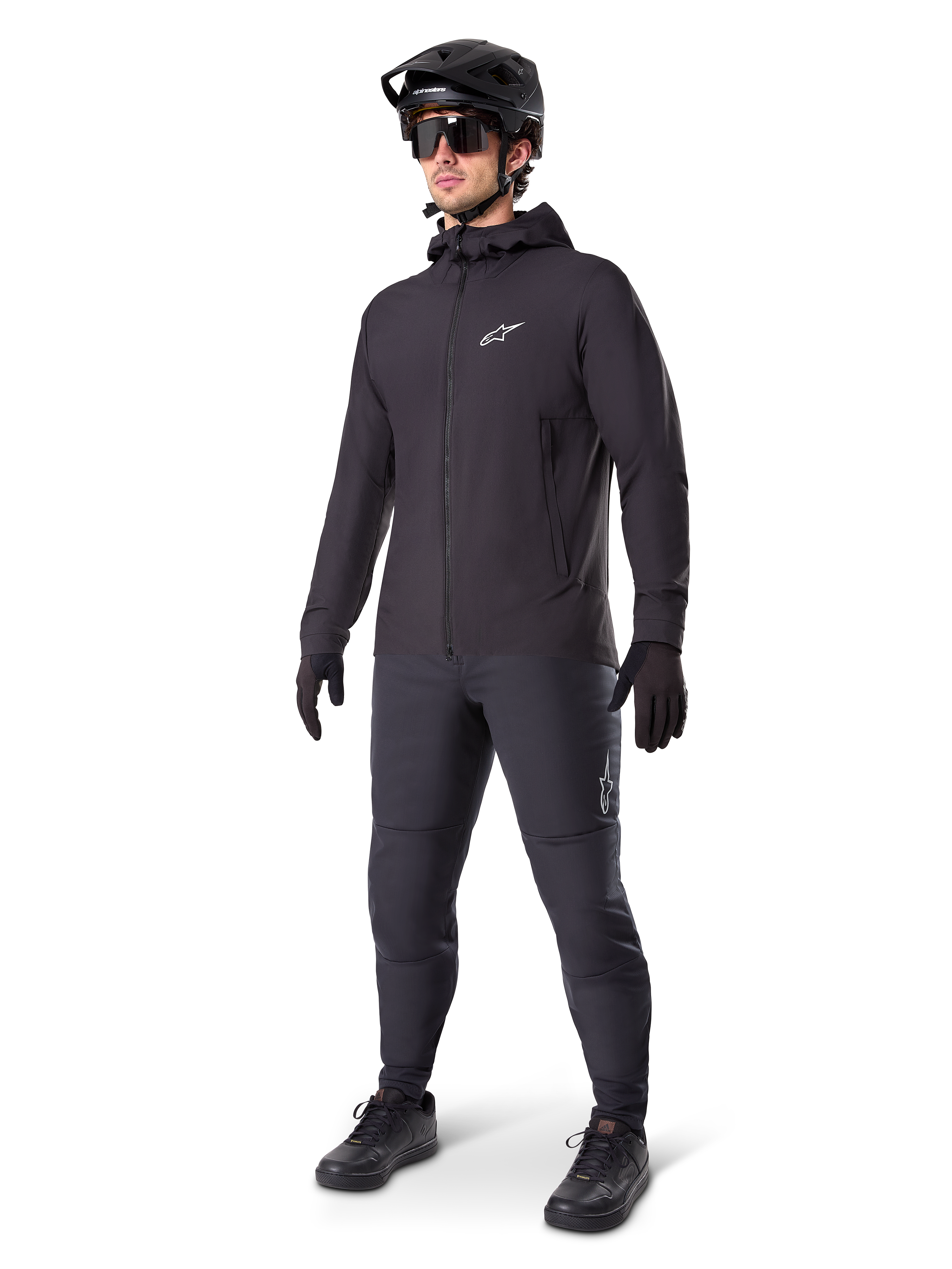 Alpinestars A-Dura Thermal Jacket w kolorze czarnym, noszona przez rowerzystę w pełnym stroju MTB, w tym czarnym kasku, okularach przeciwsłonecznych i spodniach rowerowych, lekka ocieplana kurtka z kapturem do jazdy w niskich temperaturach.