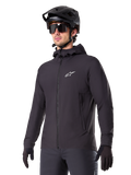 Alpinestars A-Dura Thermal Jacket, czarna kurtka rowerowa, fason z kapturem i zamkiem błyskawicznym z przodu, białe logo z gwiazdą na piersi, noszona z czarnym kaskiem MTB i rękawiczkami, sportowa odzież wyczynowa do jazdy na szlaku w zimne dni