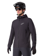 Alpinestars A-Dura Thermal Jacket, czarna kurtka rowerowa, fason z kapturem i zamkiem błyskawicznym z przodu, białe logo z gwiazdą na piersi, noszona z czarnym kaskiem MTB i rękawiczkami, sportowa odzież wyczynowa do jazdy na szlaku w zimne dni