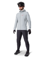 Alpinestars A-Dura Thermal Jacket w kolorze Titanium, jasnoszara rowerowa kurtka z kapturem z czerwonym logo Alpinestars na piersi, noszona przez mężczyznę w czarnym kasku MTB, okularach przeciwsłonecznych, ciemnoszarych spodniach i czarnych rękawiczkach, profesjonalny sprzęt MTB