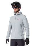 Alpinestars A-Dura Thermal Jacket, jasnoszara ocieplana kurtka rowerowa, model z kapturem i małym czerwonym logo, noszona z czarnym kaskiem i rękawiczkami, techniczna odzież na rower górski do jazdy w niskich temperaturach