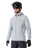 Alpinestars A-Dura Thermal Jacket, jasnoszara ocieplana kurtka rowerowa, model z kapturem i małym czerwonym logo, noszona z czarnym kaskiem i rękawiczkami, techniczna odzież na rower górski do jazdy w niskich temperaturach