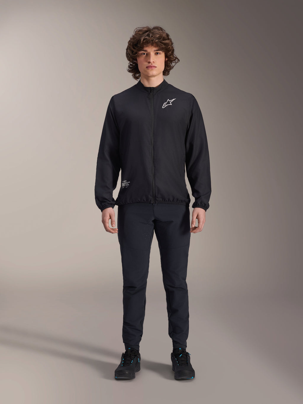 Model w czarnej kurtce Alpinestars A-Aria Windbreaker, z zamkiem na całej długości, stójką i białym logo Astars na piersi, w zestawie z czarnymi spodniami technicznymi i butami, odzież rowerowa do jazdy górskiej i użytku na zewnątrz.