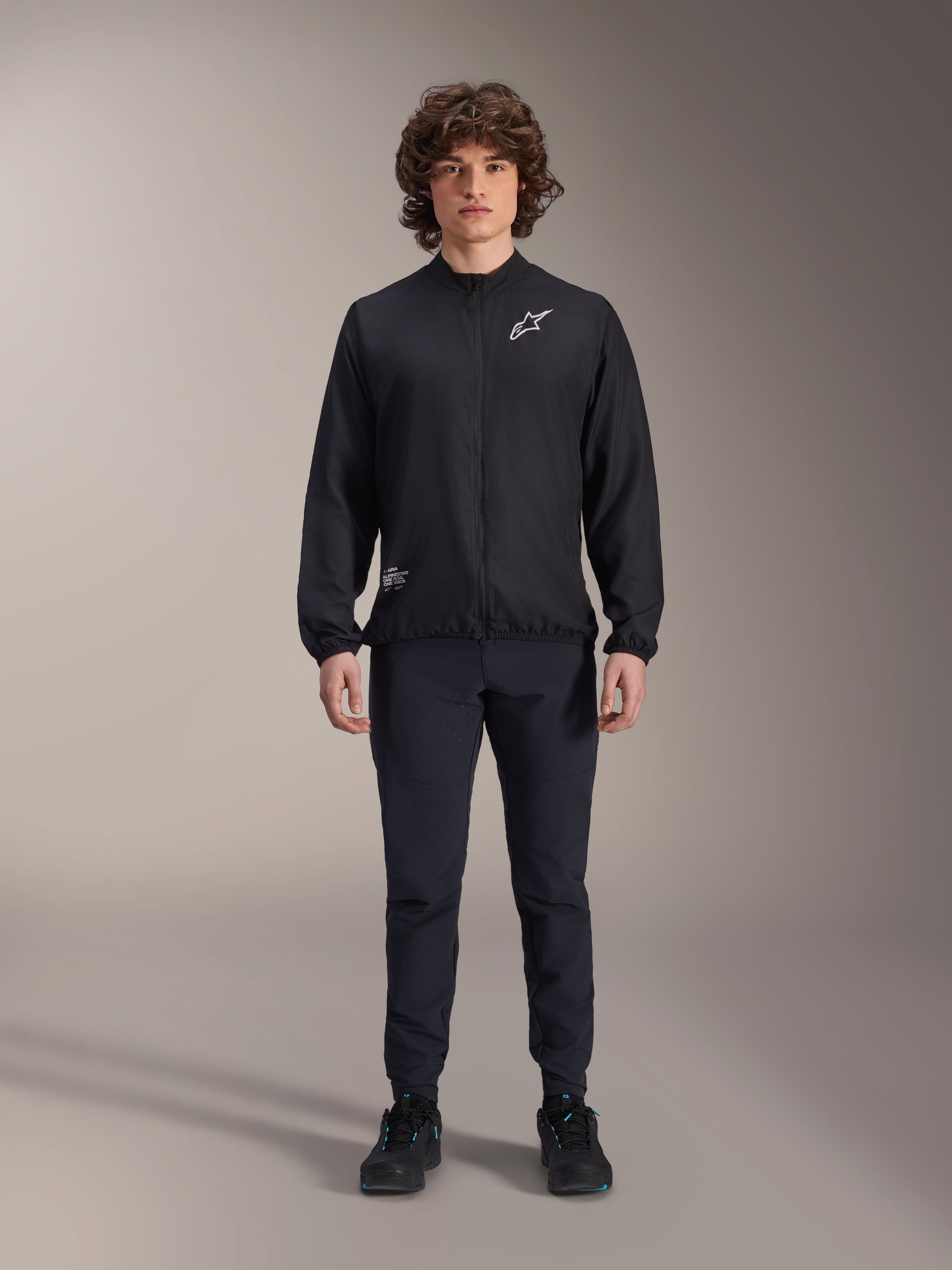Model w czarnej kurtce Alpinestars A-Aria Windbreaker, z zamkiem na całej długości, stójką i białym logo Astars na piersi, w zestawie z czarnymi spodniami technicznymi i butami, odzież rowerowa do jazdy górskiej i użytku na zewnątrz.