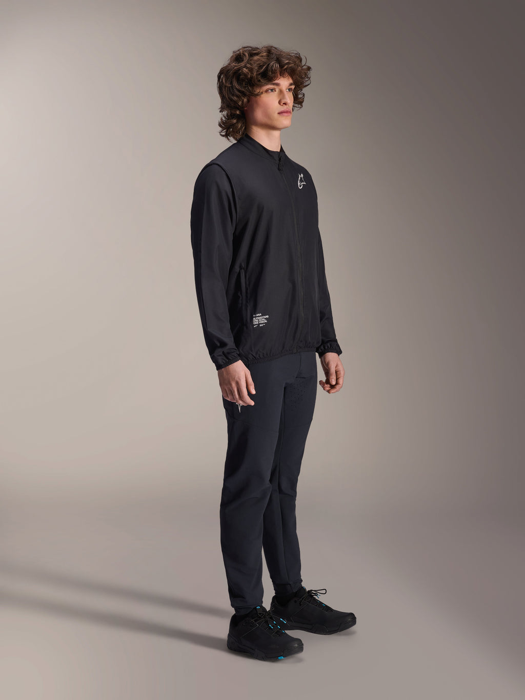 Alpinestars A-Aria Windbreaker Jacket, czarna, lekka wiatroodporna kurtka rowerowa noszona przez modela z kręconymi włosami, z białym logo Astars na piersi i brandingiem na dole po prawej stronie, zestawiona z czarnymi spodniami i butami, sportowy krój idealny do jazdy MTB i gravelowej