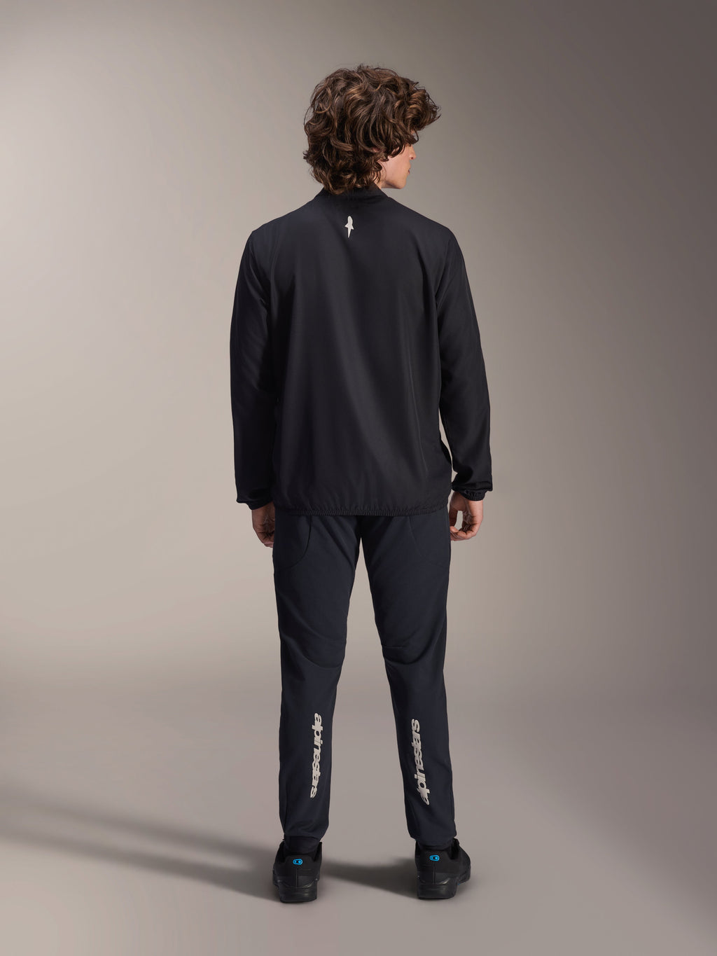 Alpinestars A-Aria Windbreaker Jacket, czarna, widok z tyłu, lekka wiatroodporna kurtka rowerowa z małym białym logo w górnej części pleców, noszona z dopasowanymi czarnymi spodniami z pionowym napisem Alpinestars na łydkach, profesjonalna odzież MTB