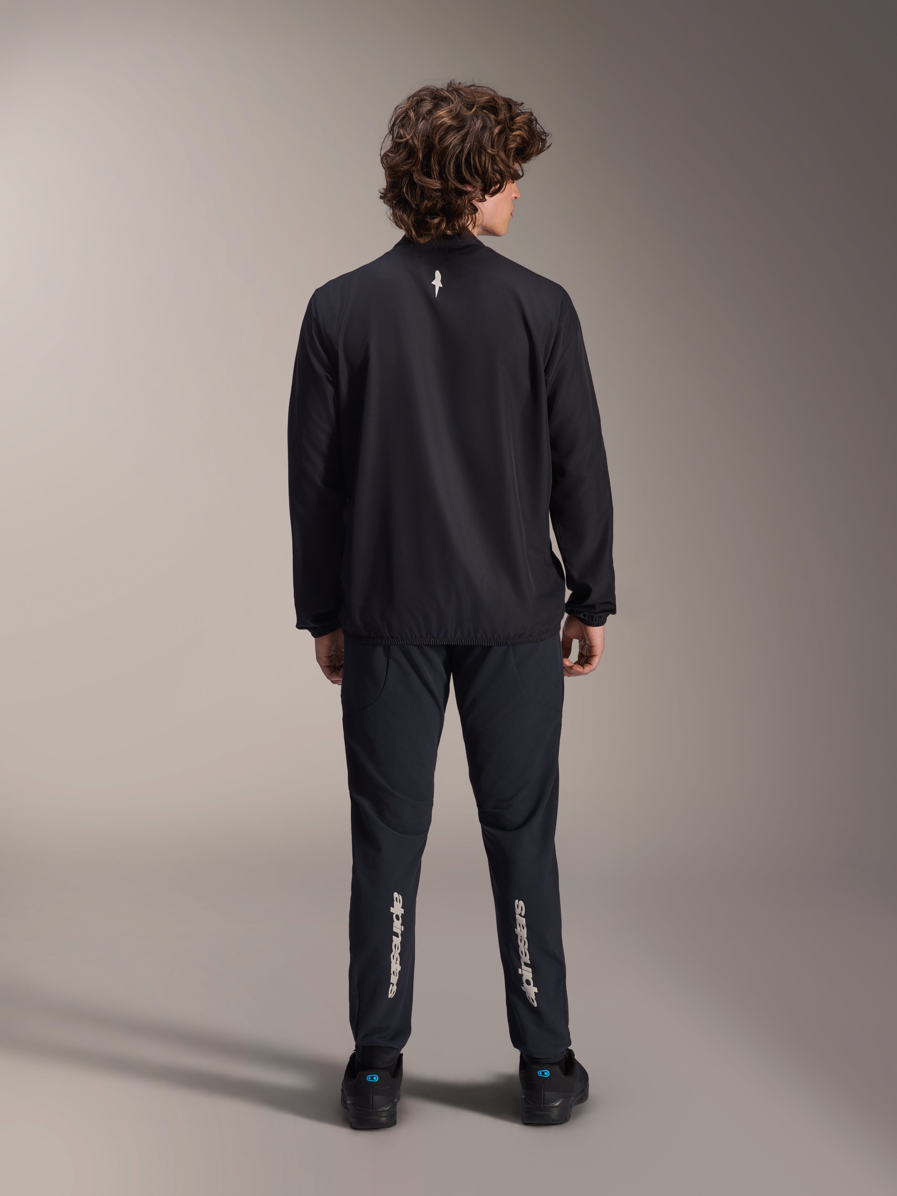 Alpinestars A-Aria Windbreaker Jacket, czarna, widok z tyłu, lekka wiatroodporna kurtka rowerowa z małym białym logo w górnej części pleców, noszona z dopasowanymi czarnymi spodniami z pionowym napisem Alpinestars na łydkach, profesjonalna odzież MTB