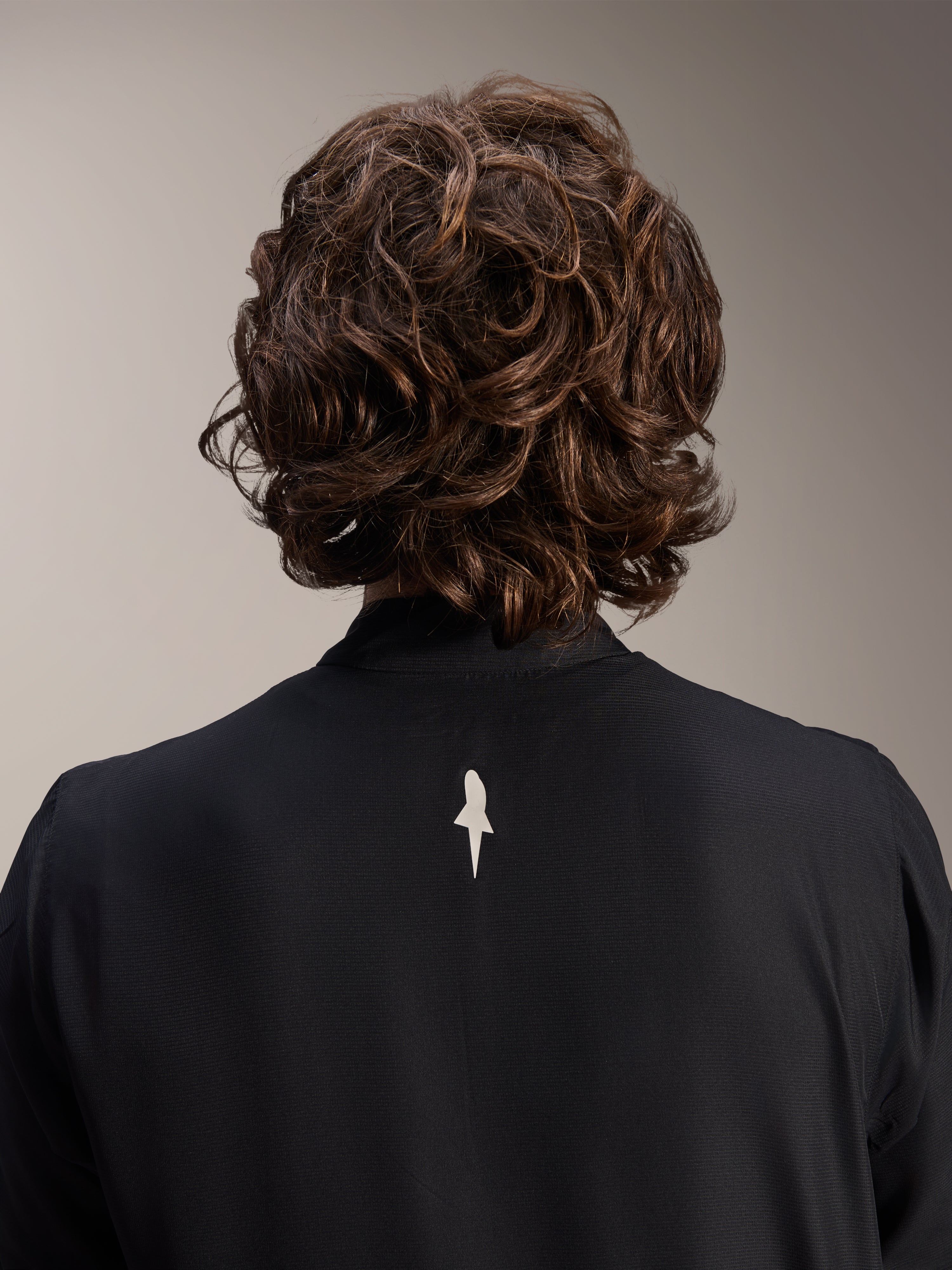 Kurtka Alpinestars A-Aria Windbreaker, czarna, zbliżenie z tyłu na górną część ramion i kołnierz, małe białe logo na środku pleców, model z brązowymi falowanymi włosami, minimalistyczna wyczynowa odzież rowerowa