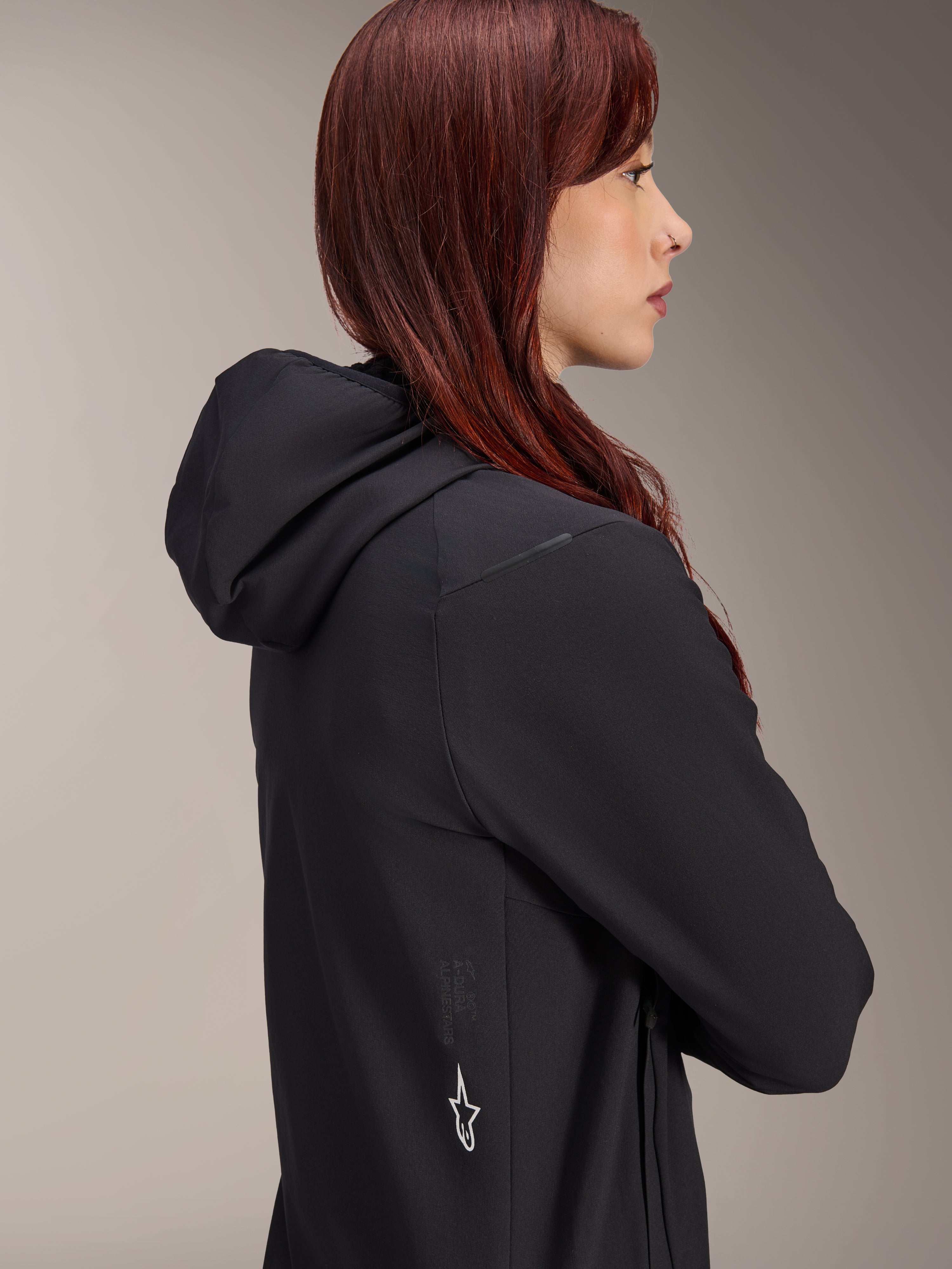 Kurtka Alpinestars Stella A-Dura Thermal, damska kurtka rowerowa, czarna, profil kobiety o rudych włosach ubranej w kurtkę, zintegrowany kaptur i białe logo gwiazdy Alpinestars z boku, lekka ocieplana odzież termiczna do MTB
