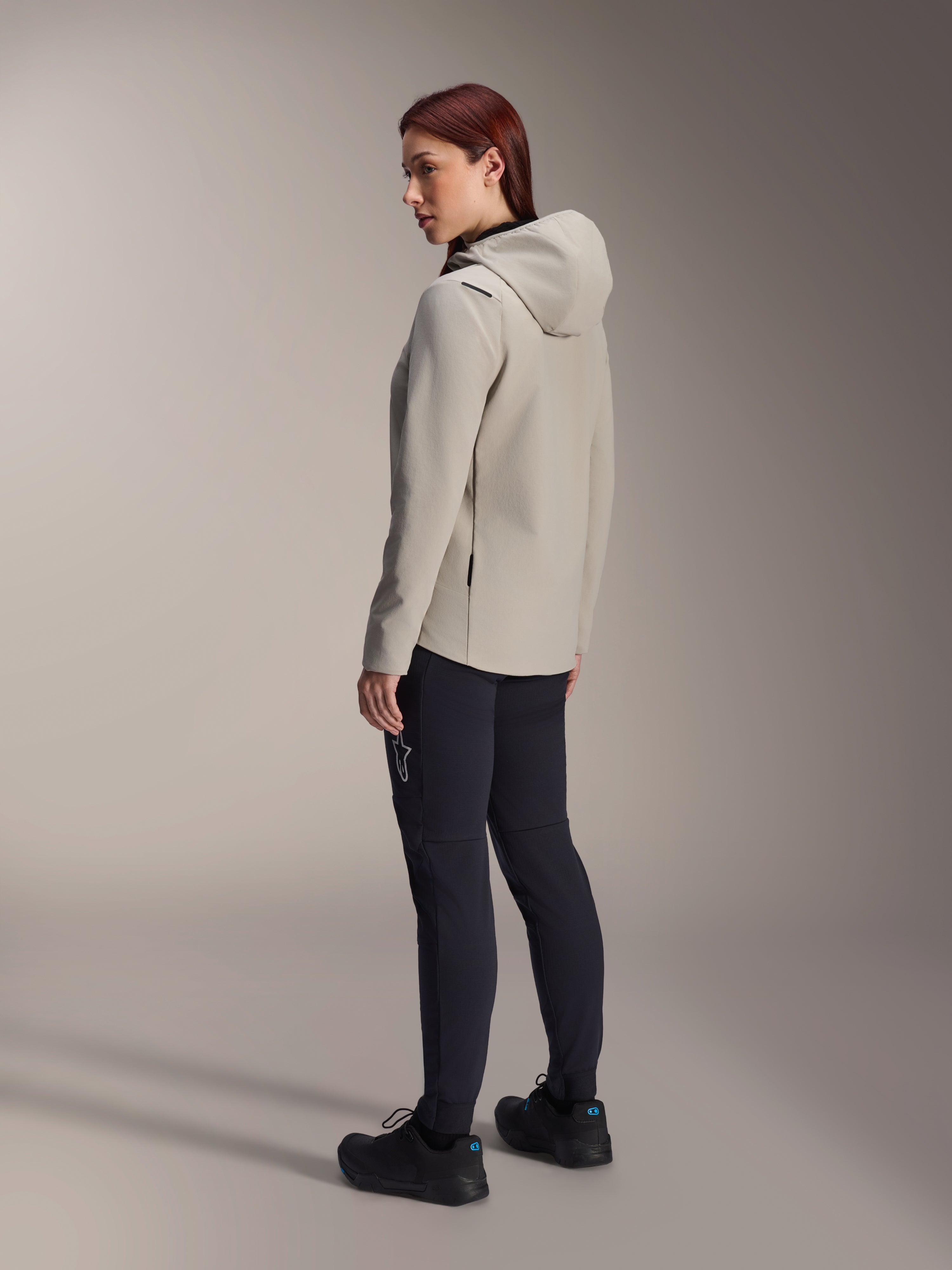 Alpinestars Stella A-Dura Thermal Jacket, szara ocieplana kurtka rowerowa z kapturem, widok z tyłu pod kątem trzech czwartych na kobietę w czarnych spodniach MTB, ujęcie studyjne, regularny krój i detale kieszeni