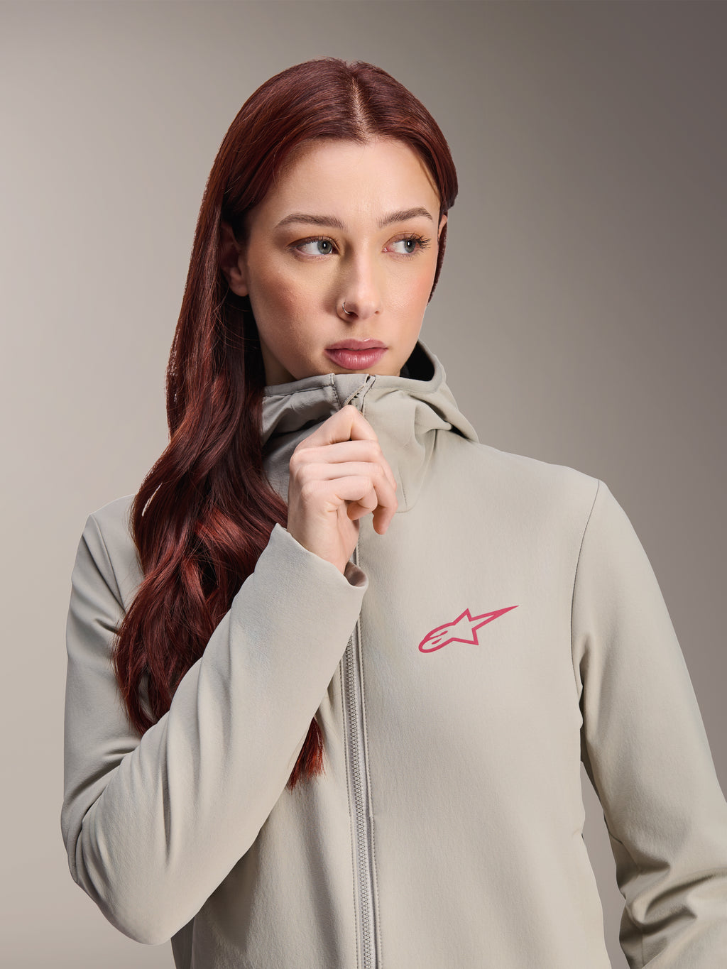 Damska kurtka termiczna Alpinestars Stella A-Dura, kolor szary Titanium z różowym logo gwiazdy Alpinestars, modelka z długimi rudymi włosami zapinająca kurtkę z kapturem, lekka ocieplana odzież rowerowa na zimną pogodę