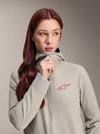 Damska kurtka termiczna Alpinestars Stella A-Dura, kolor szary Titanium z różowym logo gwiazdy Alpinestars, modelka z długimi rudymi włosami zapinająca kurtkę z kapturem, lekka ocieplana odzież rowerowa na zimną pogodę