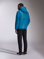 Alpinestars Speeder Chromium Windbreak Jacket, niebieska kurtka przeciwwiatrowa z kapturem, widok z tyłu z czarnym napisem logo Alpinestars na plecach, noszona z czarnymi spodniami i czarno-białymi tenisówkami, casualowy streetwear inspirowany motorsportem