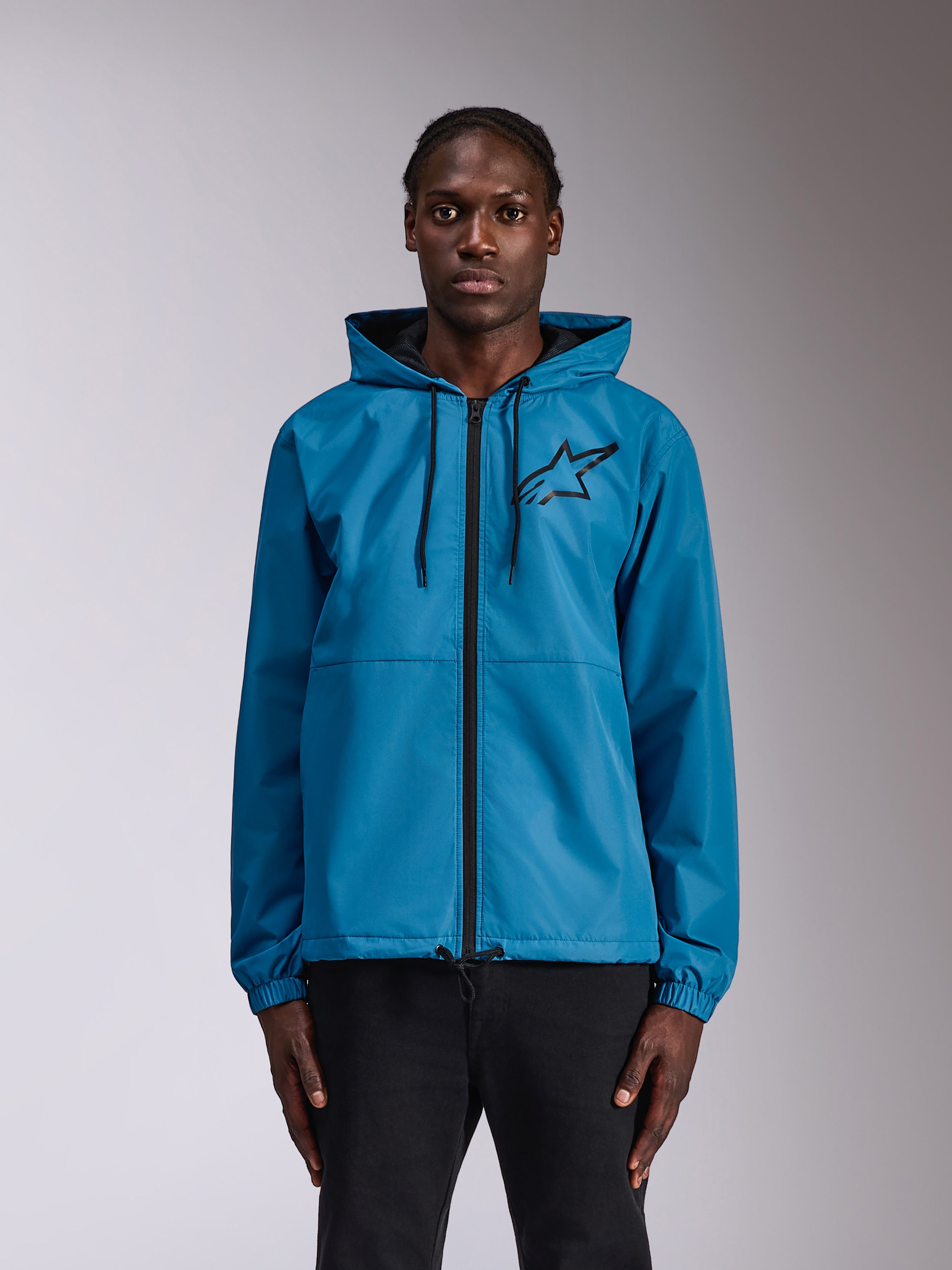 Alpinestars Speeder Chromium Windbreak Jacket, niebieska kurtka przeciwwiatrowa z kapturem, czarne logo Astars na piersi, pełny zamek błyskawiczny, elastyczne mankiety i ściągacz przy kapturze, prezentowana przez modela jako sportowa odzież codzienna