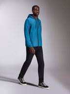 Alpinestars Speeder Chromium Windbreak Jacket, niebieska wiatrówka z kapturem prezentowana przez modela, posiada czarny zamek z przodu i logo na piersi, w zestawieniu z czarnymi spodniami i wysokimi butami sportowymi, codzienna odzież sportowa inspirowana motorsportem