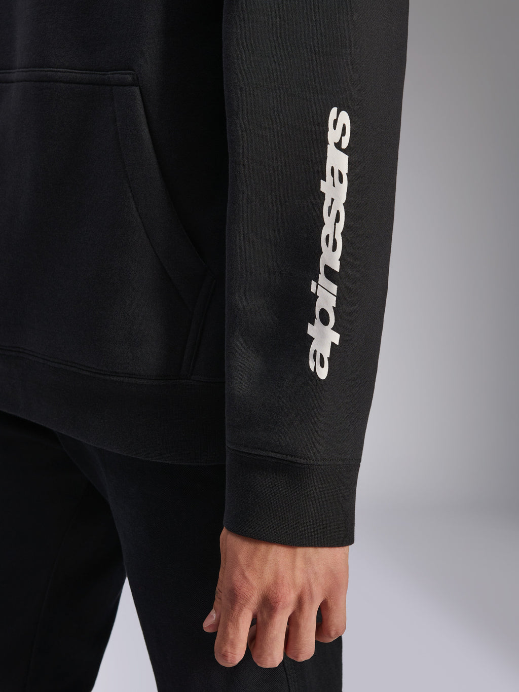 Alpinestars Harken Hoodie, czarna, zbliżenie na rękaw z pionowym białym logotypem, ściągacz przy mankiecie i detal kieszeni typu kangurka, codzienna polarowa odzież sportowa