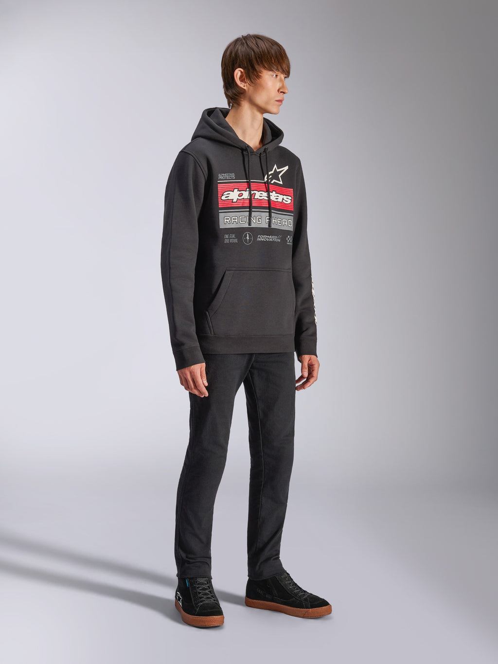 Alpinestars Harken Hoodie, czarna bluza polarowa z kapturem wkładana przez głowę, z dużą grafiką na piersi, czerwonym napisem Alpinestars i logo gwiazdy, kieszeń kangurka, noszona przez modela z czarnymi spodniami i sneakersami, casualowa odzież streetwearowa